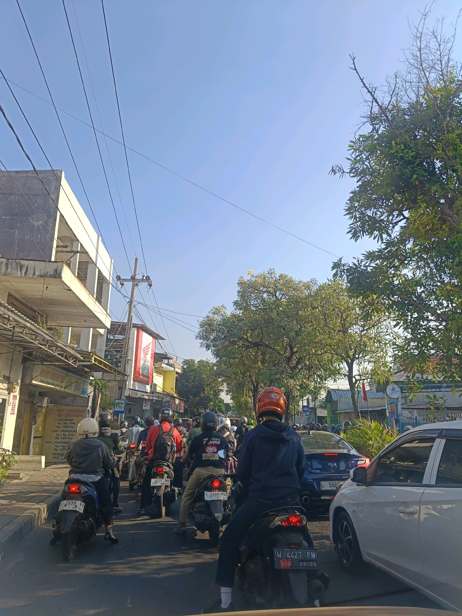 The image depicts a busy street scene in Surabaya, Indonesia, characterized by a traffic jam of motorcycles and cars under a clear blue sky. The perspective is from a vehicle in traffic, looking forward.
In the foreground, several motorcycles are visible, with riders wearing helmets of various colors, including orange, white, and black. Some riders are looking ahead, while others appear to be stationary. One person in the center is wearing a dark blue hoodie, and another to their left wears a grey jacket. A white car is parked to the right, and a blue Subaru car is in front of it, with its license plate partially visible. The license plates on the motorcycles show Indonesian alphanumeric codes.
The environment includes buildings on the left side of the street, with a multi-story structure featuring balconies and a gray facade. A utility pole with numerous wires is also present. Advertisements and signage are visible on the buildings, including a large red and white banner with the Honda logo. Trees with green foliage line the right side of the street, with some bare branches visible. The road surface is asphalt, and a sidewalk is present on the left. The time of day appears to be morning or late afternoon, given the sunlight and shadows.