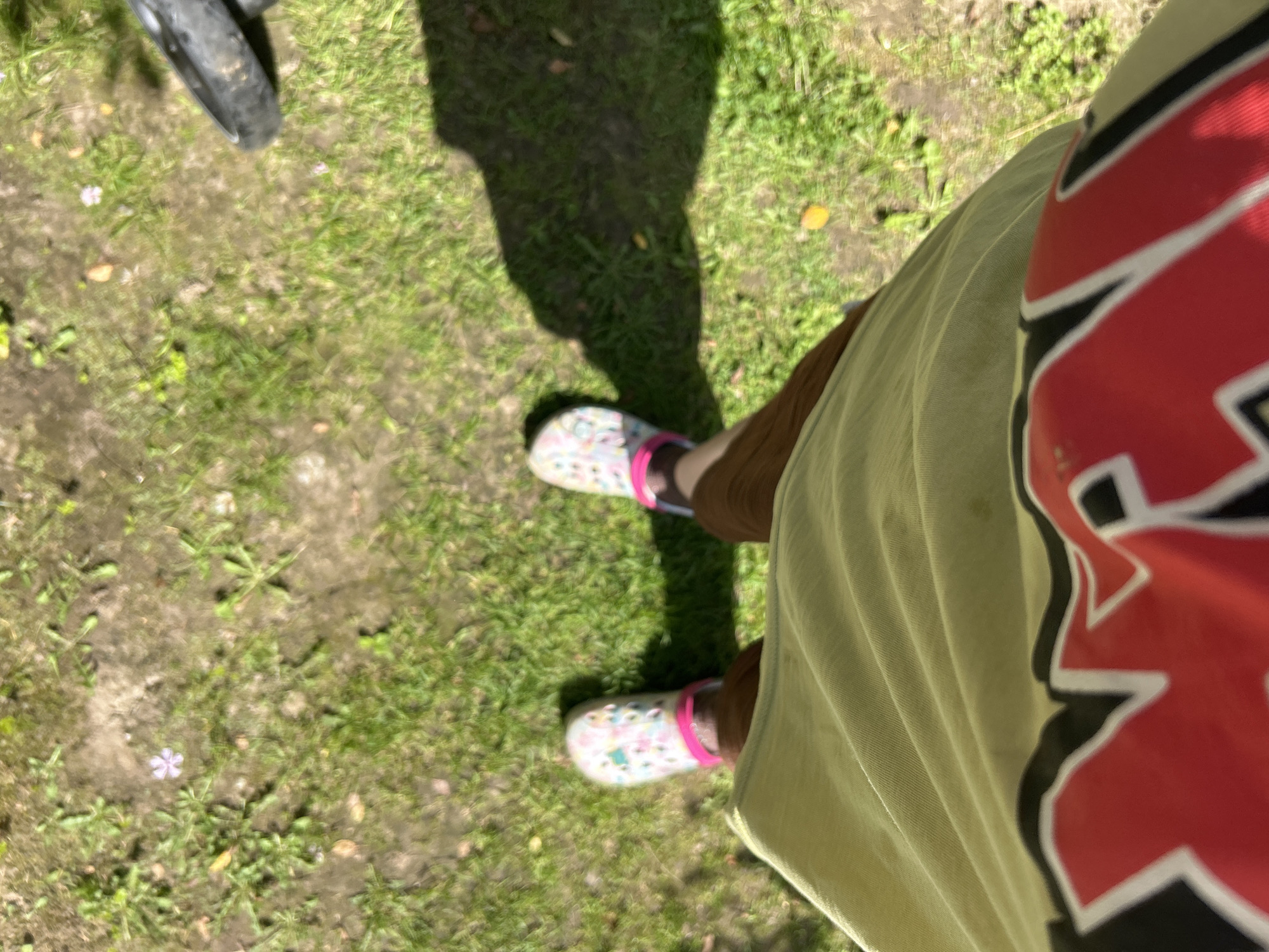 A first-person perspective shot shows a person standing on a grassy area with patches of dirt. The person is wearing patterned Crocs with pink straps and brown pants. A portion of a light olive-green garment, possibly a t-shirt, is visible at the bottom of the frame, with a bold red and white graphic design on it. A shadow of the person stretches across the ground to the right. In the background, a black tire and part of a bicycle are visible. A small purple flower is on the ground near the person's left foot. The setting appears to be outdoors, possibly a park or yard.