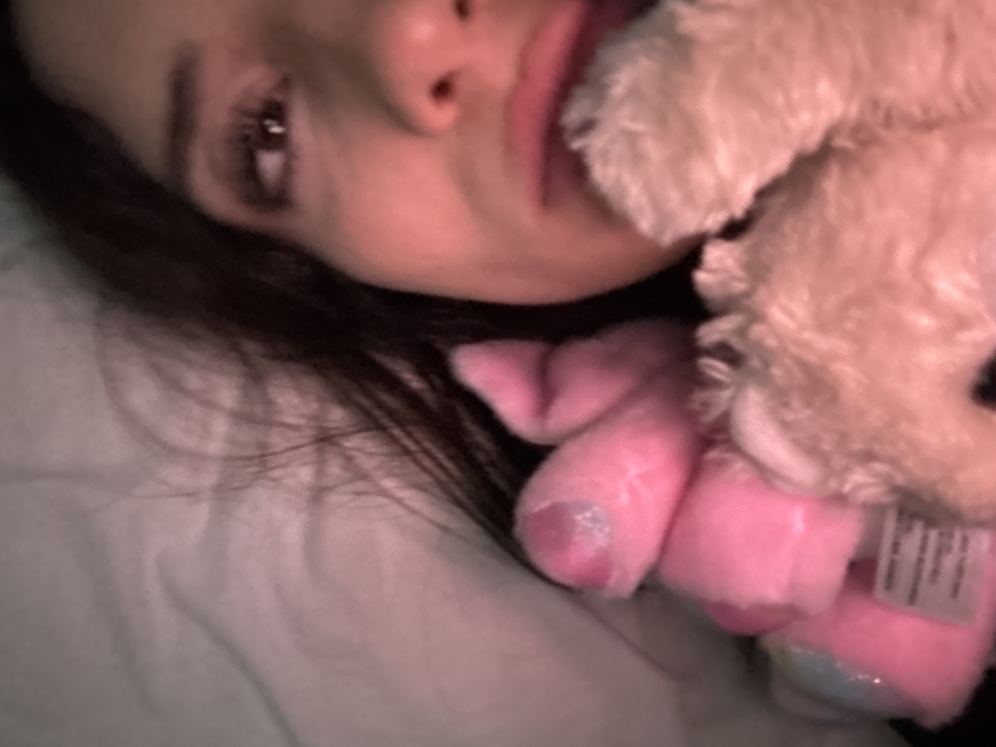 A person with dark hair is lying in a bed with white sheets, holding a plush toy. The toy is light pink and cream-colored, with visible tags. The person's face is visible, with an eye open and lips partially pursed. The location is unknown, and there is no information to indicate the city of Buenos Aires, Argentina.