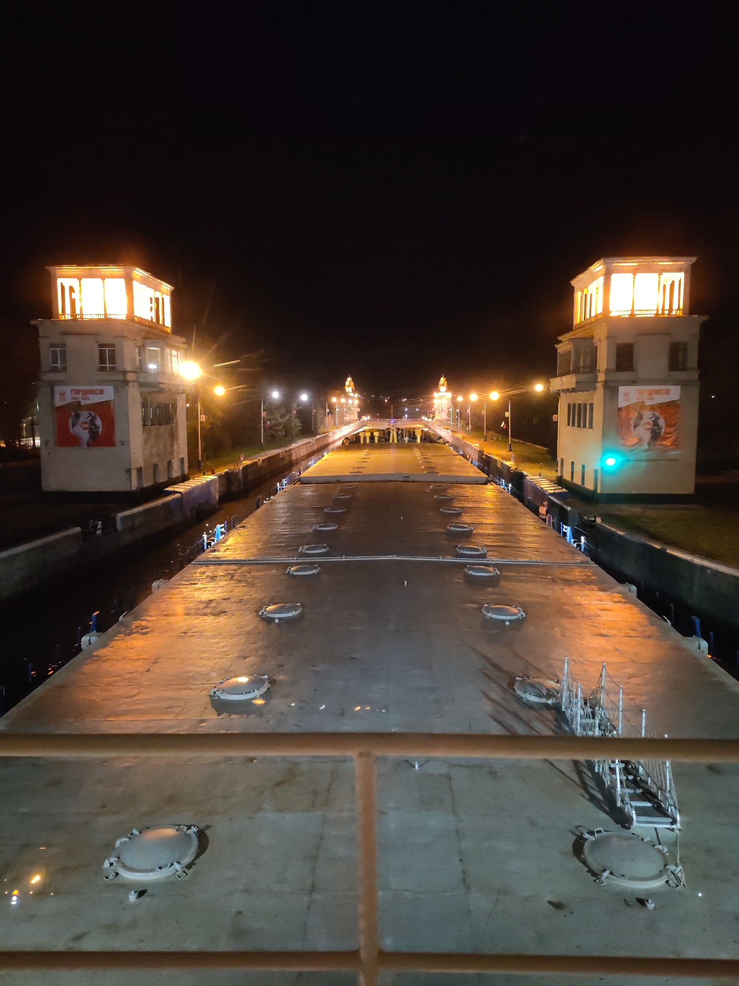 This image captures the scene at Lock No. 1 of the Moscow Canal in Yakhroma, Russia at night. The canal stretches between two imposing, lighted towers. Each tower has large posters. The canal's surface, punctuated by circular hatches, reflects the ambient light. In the foreground, a railing suggests the viewpoint is from a ship. Street lamps illuminate the route along the canal, disappearing into the distance. The sky is dark, contrasting with the bright lights.