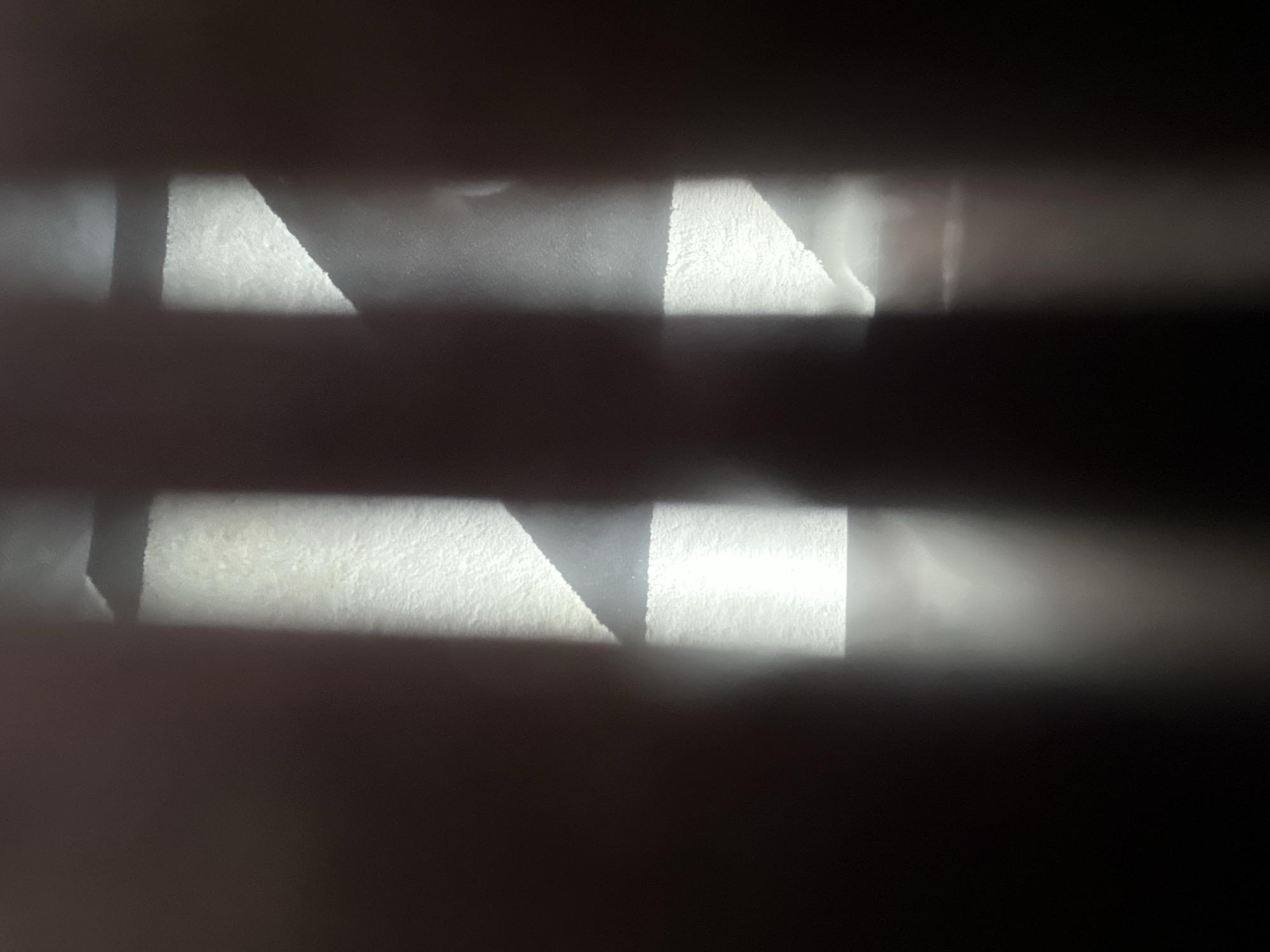 This is a close-up, horizontally oriented photograph of what appears to be a surface with a textured, light-colored material. The surface is divided into horizontal bands by dark, out-of-focus areas. Within these bands, there are abstract geometric shapes formed by the interplay of light and shadow. One prominent shape is a triangle with a rough, granular texture, illuminated by a bright light source. This triangle is bordered by a darker, smoother area that transitions into a bright, almost blown-out section, suggesting a strong light source or reflection. The lighting creates a high-contrast scene with deep shadows and bright highlights. The overall impression is abstract and textural. There is no discernible location context beyond the provided metadata suggesting Belgrade, Serbia, and no text is visible. The time of day and weather are not ascertainable from the image itself.