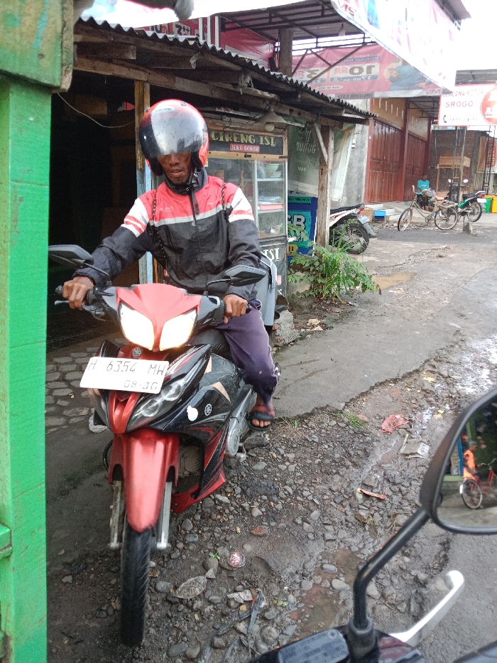 A person wearing a helmet, jacket, and pants sits on a red and black motorcycle, facing the viewer. The motorcycle has a license plate that reads "H 6354 MH". The motorcycle is on a street with a rough surface. Behind the person are buildings, one of which has a sign that says "Goreng ISI". In the foreground, a motorcycle mirror is visible. There is also another motorcycle, as well as bicycles, in the background.