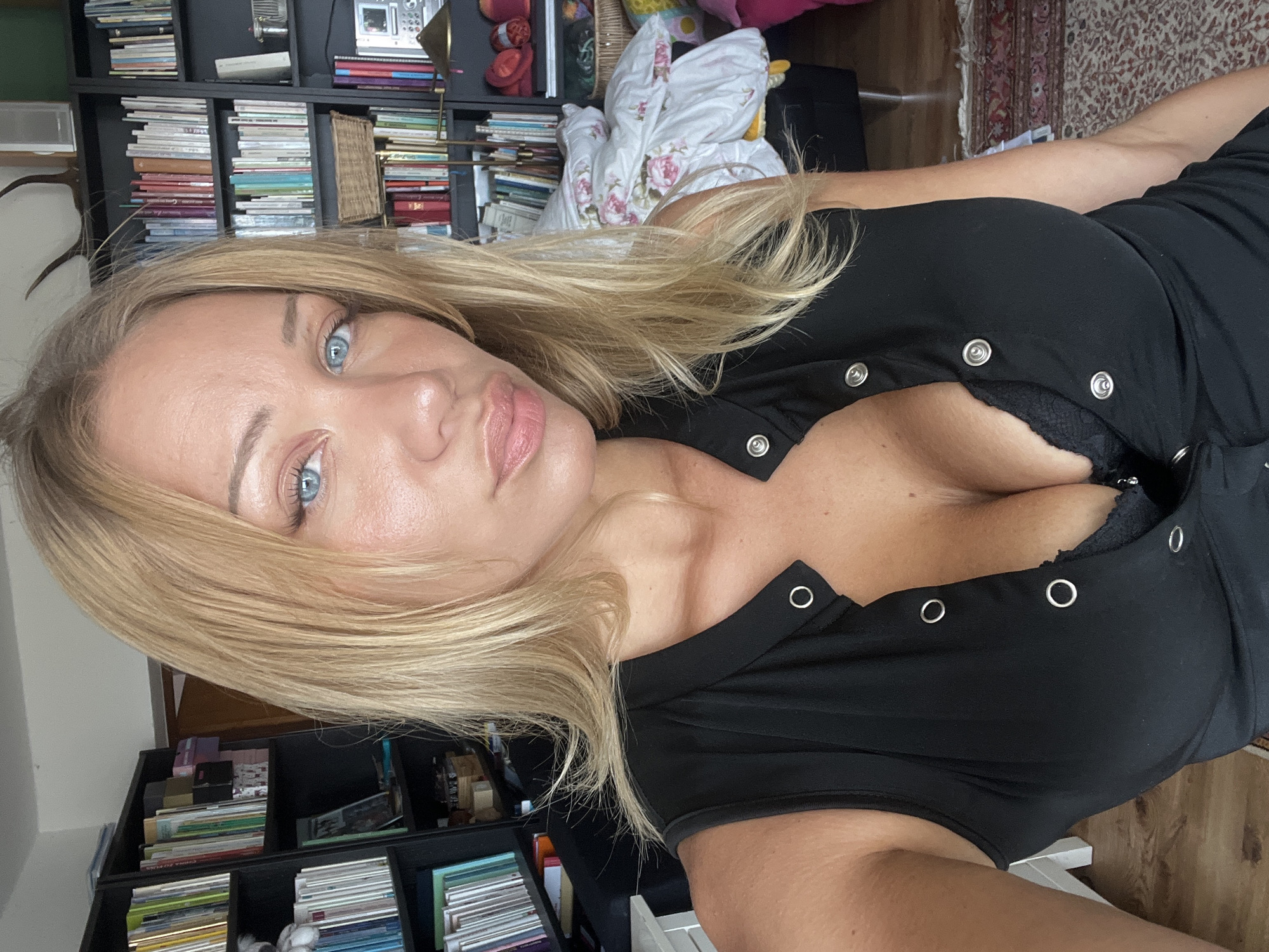 A fair-skinned woman with blonde hair and blue eyes is pictured in a selfie. She is wearing a black, sleeveless top with silver snaps and a black lace bra. Her hair is slightly windswept, and she is looking directly at the camera. Behind her, bookshelves filled with books are visible. The lighting suggests an indoor environment, possibly a home, with natural light illuminating the scene. The specific location in Mühldorf a. Inn, Germany is not discernible from the image alone.