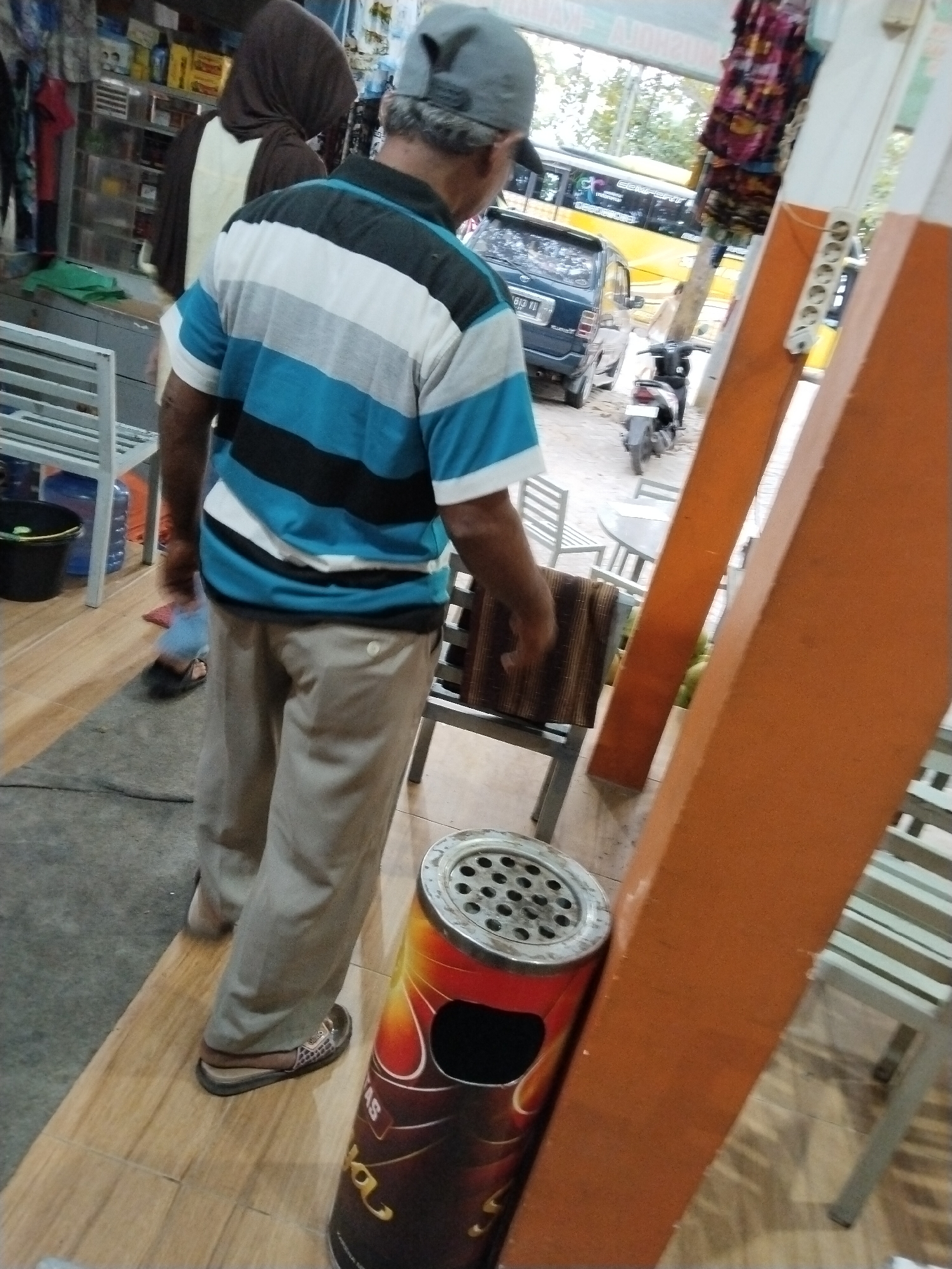 The image depicts a street scene in Prigi, Indonesia, with a focus on a man standing near an outdoor seating area. The man, wearing a gray baseball cap and a striped polo shirt with blue, black, and white bands, is facing away from the camera. He is also wearing light-colored trousers and sandals. To his left, another person is partially visible, wearing a dark headscarf and a light-colored garment. Behind the man, a dark blue car is parked, and further back, a yellow bus is visible, suggesting a public thoroughfare. In the foreground, a cylindrical ashtray with a metal top and a decorative, flame-like pattern on its body is prominent. The environment includes wooden flooring, what appears to be metal chairs and tables, and some retail displays in the background with various goods. The overall impression is of a commercial or public space in a tropical setting.
