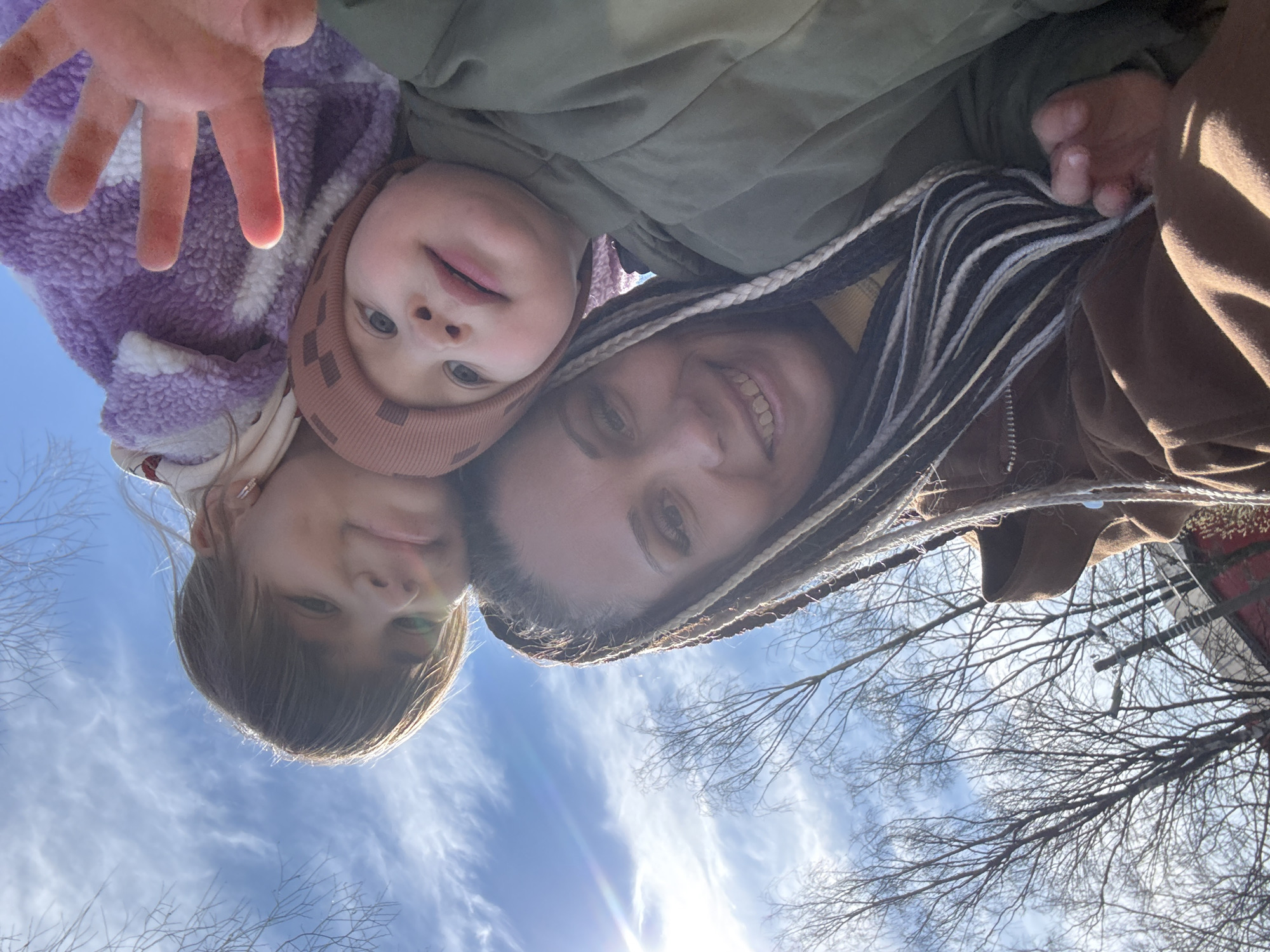 A selfie shows a woman with two children outdoors against a backdrop of a blue sky with wispy clouds and bare tree branches. The photograph is taken from a low angle, looking upwards. The woman is smiling, has light skin, and braided hair. One child wears a pink knit cap and a purple, textured jacket and extends their hand towards the camera. The other child has shoulder-length brown hair. The location is inferred to be Yaroslavl', Russia, based on the prompt, but there are no explicit indicators of the city visible in the frame.
