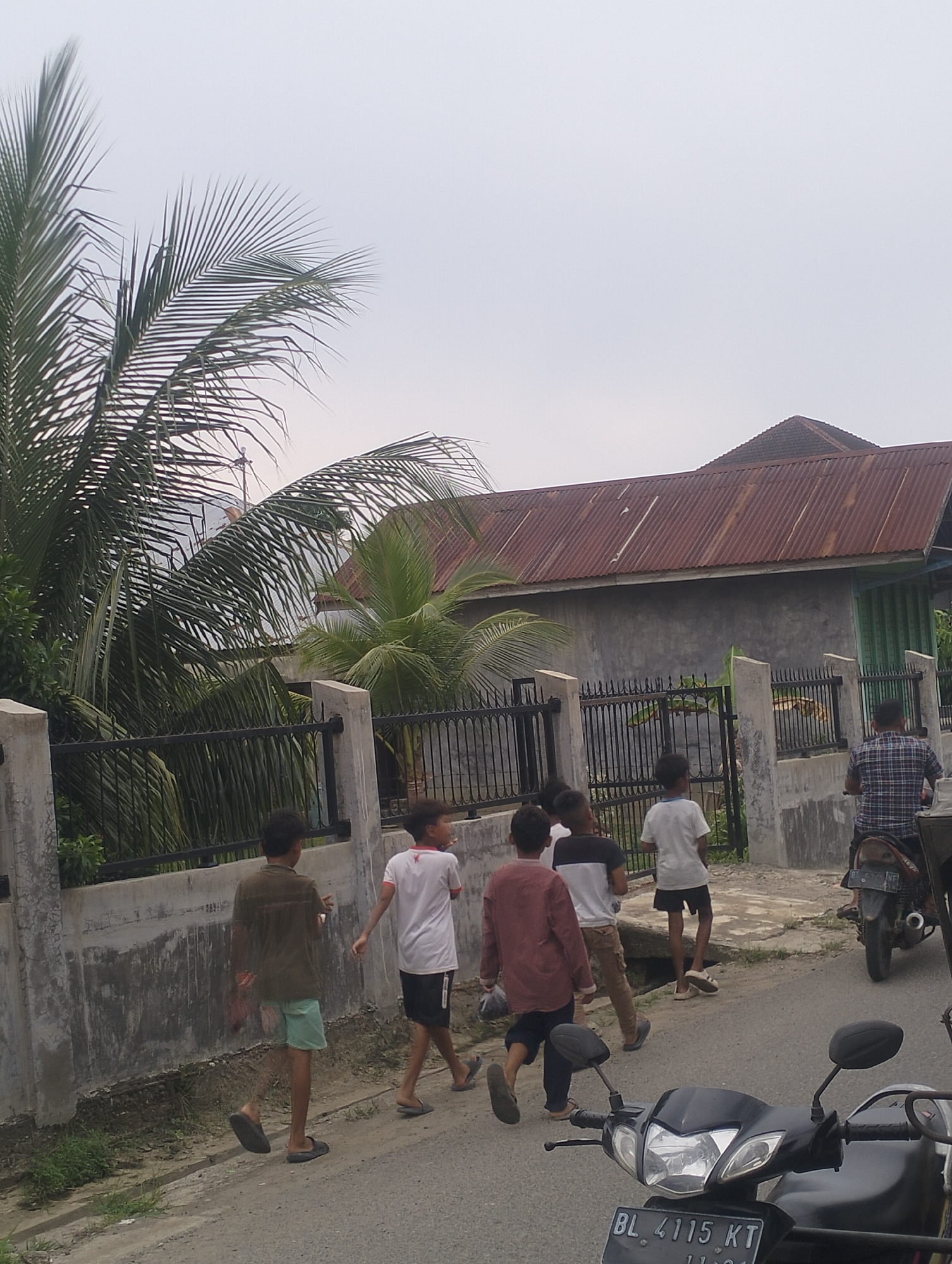 In Lhokseumawe, Indonesia, on a paved road next to a concrete wall with a black metal fence, a group of young boys walk away from the camera. The boy on the far left wears a brown shirt and light blue shorts. Next to him, a boy in a white t-shirt and black shorts walks. A third boy in a maroon long-sleeved shirt and dark shorts is partially obscured. Another boy in a white t-shirt and khaki pants is visible further back. A man on a motorcycle, wearing a blue and white checkered shirt, rides away from the group on the road. In the background, a house with a rusty corrugated metal roof and gray walls is visible, partially surrounded by palm trees and other vegetation. The sky is overcast.
