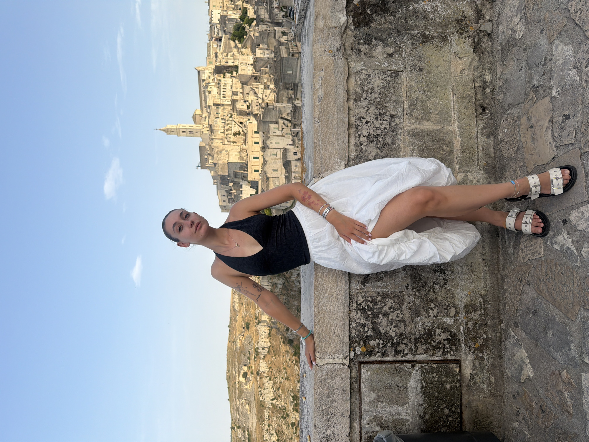 A young woman is posing in front of the ancient city of Matera, Italy. She is leaning against a stone wall, with her left arm resting on it. She is wearing a black halter top and a flowing white skirt, along with white sandals. Her legs are crossed and her right leg is slightly lifted, revealing her knee. She has several tattoos on her arms. In the background, the city of Matera, a UNESCO World Heritage site, is visible with its distinctive cave dwellings and architecture, built into the side of a hill. The sky above is a clear blue with a few wispy clouds. The foreground consists of a stone walkway and the stone wall the woman is leaning on.