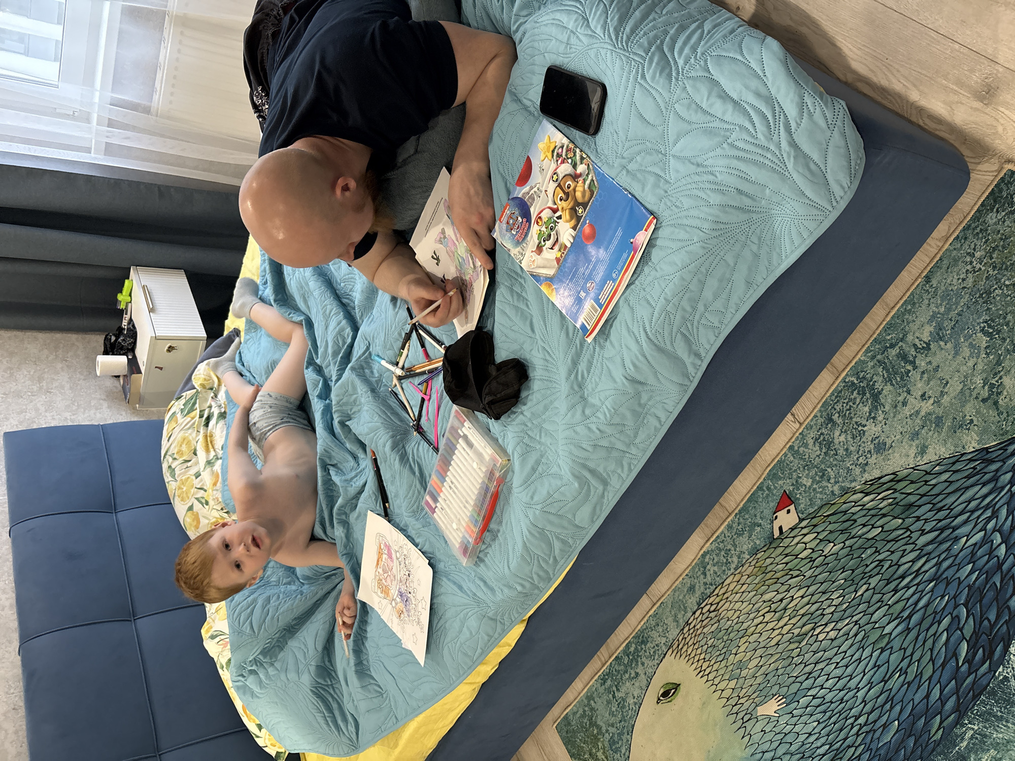 A man and a child are lying on a bed. The man is bald, wearing a dark blue t-shirt and grey pants, and he is coloring a picture with a pencil. The child is fair-skinned with reddish hair, wearing grey shorts, and is looking at the camera while also holding a coloring page. The bed has a blue and yellow patterned bedspread. A black mobile phone, a Paw Patrol coloring book, a set of colored markers, and a black cap are on the bed. A rug with an image of a fish with a house on its back is on the floor. The location is in a room with light-colored walls and wooden floors. The city is Izhevsk, Russia.