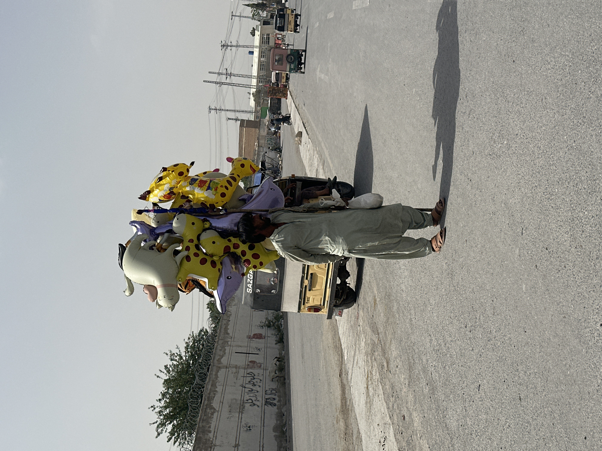 A street vendor walks along a paved road in Quetta City Tehsil, Pakistan, pushing a motorized cart laden with inflated animal-shaped balloons. The vendor, dressed in a light-colored shalwar kameez and sandals, holds the cart steady as he walks along the road. The balloons are various shapes and colors including yellow giraffes and a white cat. The cart has a "SAZGA" logo on its side. The road is bordered by a concrete wall topped with barbed wire. Overhead power lines stretch across the sky. In the distance, vehicles are seen moving along the road. The lighting suggests it's daytime.