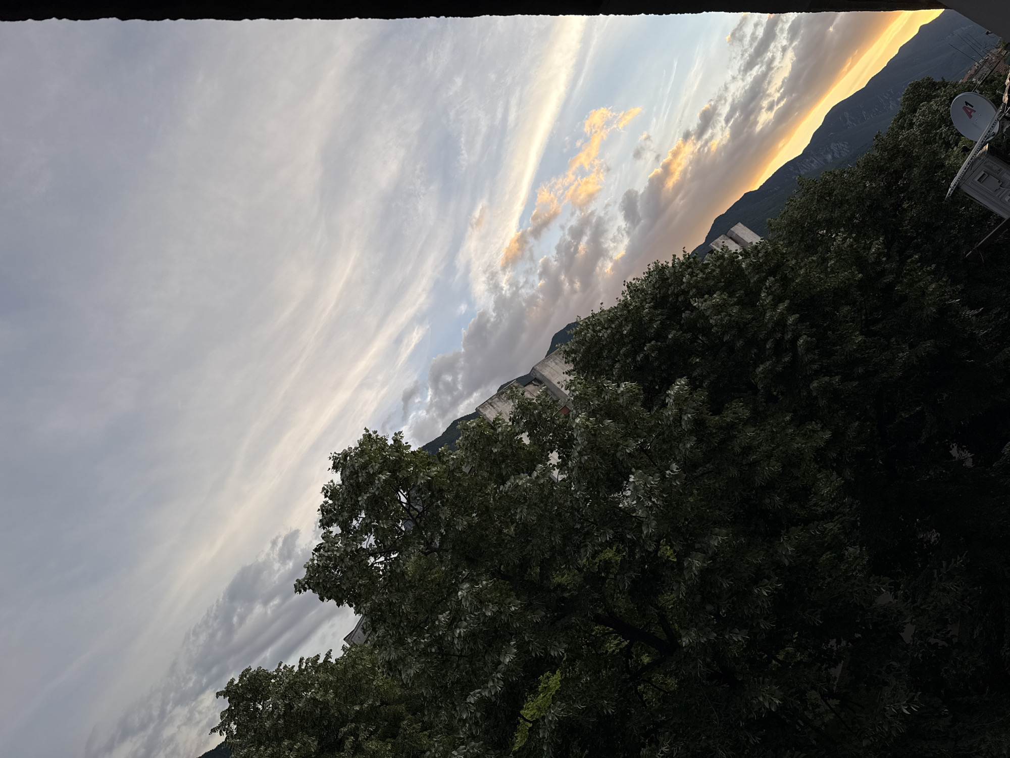 The image shows a tilted view of a landscape, likely at sunset or sunrise. In the foreground, on the left side, is a dense mass of green foliage from trees, partially obscuring buildings. The trees are lush and leafy, with some lighter green leaves catching the light. Behind the trees, a steep, dark mountain range dominates the right side of the frame, its slopes falling away towards the horizon. The sky above is a mix of pale blue, white, and soft orange and yellow hues, indicating the presence of clouds and the setting or rising sun. Wispy cirrus clouds are visible, streaked across the sky. A sliver of a dark, irregular edge runs vertically along the far right of the image, possibly a frame or a structural element. The overall impression is one of a natural scene with human elements subtly present. No specific landmarks or details are identifiable to confirm it is Vratsa, Bulgaria.