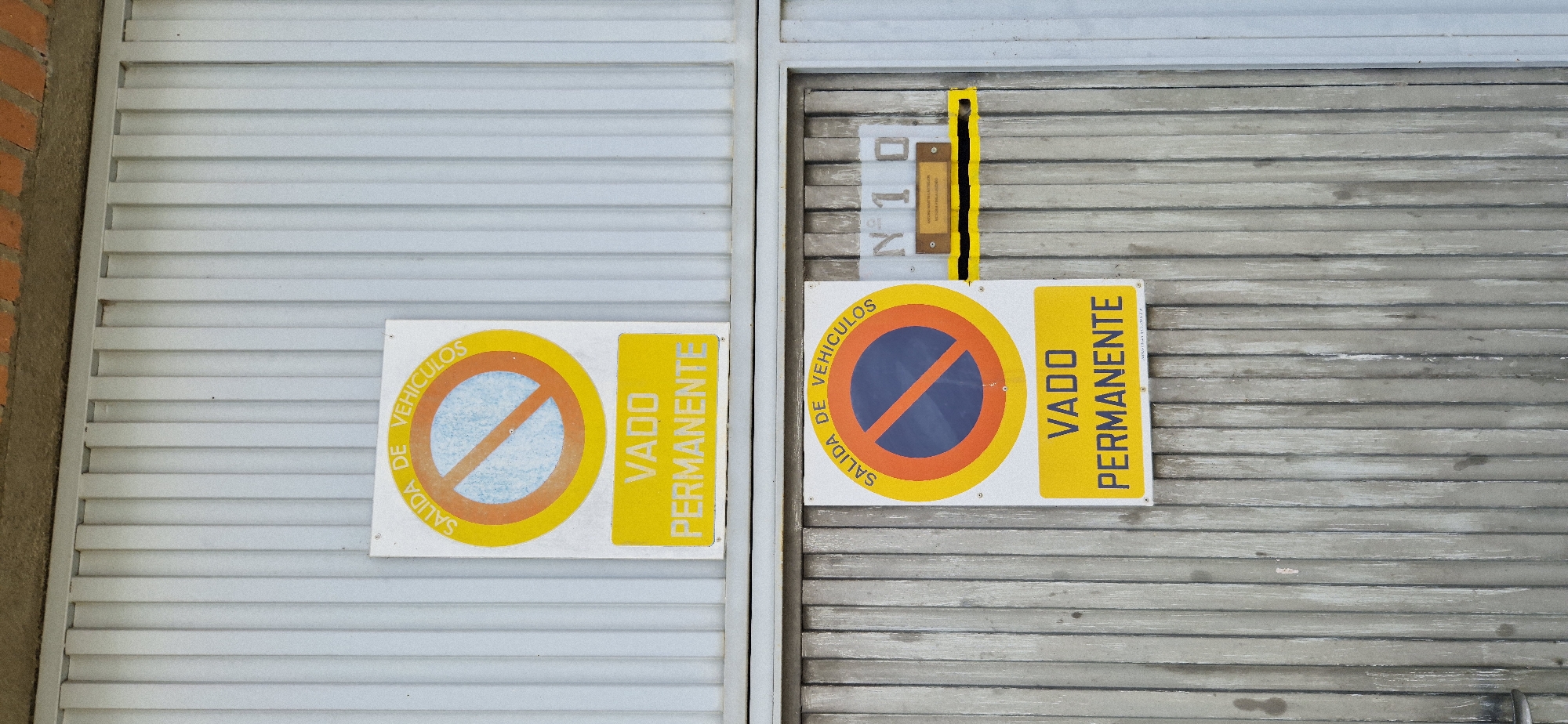 The image shows two signs on a closed garage door. The garage door on the left is made of light gray corrugated metal, and the door on the right is made of darker gray corrugated metal. Both signs are rectangular and display a universal "no parking" symbol, which is a red circle with a red diagonal line through it, superimposed on a blue area. To the right of the symbol, on a yellow background, is the text "VADO PERMANENTE," which translates to "Permanent passage" or "Permanent access" in Spanish. The text "SALIDA DE VEHICULOS" (Vehicle exit) curves around the left side of the "no parking" symbol.
The sign on the left is white with a yellow border around the "no parking" symbol and a yellow rectangular section for the text. The sign on the right is similar in design but appears slightly larger and is mounted on the darker gray garage door. Above the right sign, attached to a vertical yellow strip, is a small plaque with the inscription "N 10." There are also some very faint markings on the right sign that are difficult to decipher. The overall scene suggests a street or a private property entrance where parking is prohibited to ensure unobstructed passage. The lighting suggests it is daytime, and there are no obvious weather-related elements.