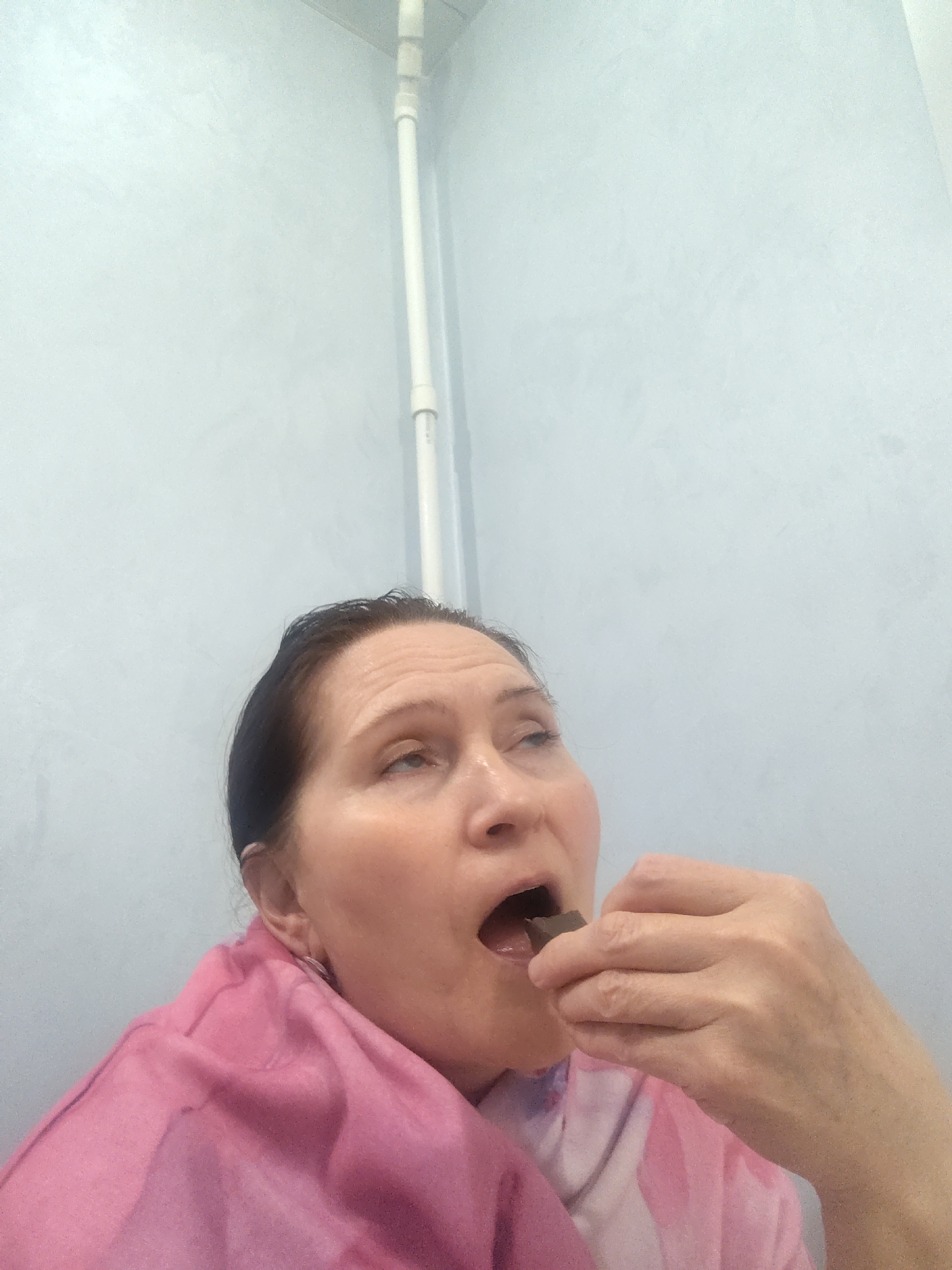 A person is in the process of eating a small, dark-colored piece of what appears to be chocolate. The individual has short dark hair, light skin, and a pink shawl or garment draped around their shoulders. The background is a light blue wall with a vertical white pipe running along it. There is no information to connect this image to the city of Neftekamsk, Russia.