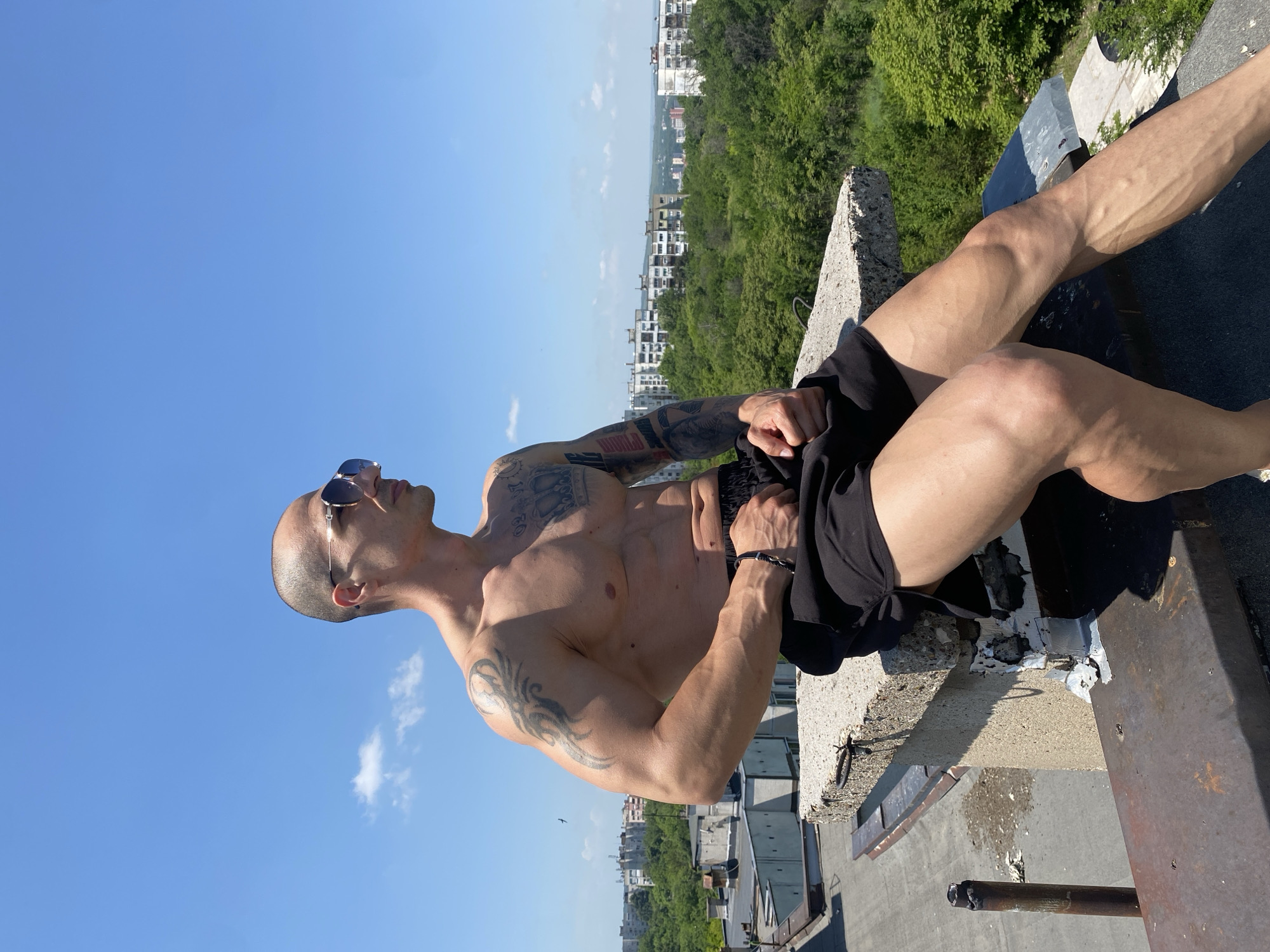 A muscular man with a shaved head and sunglasses is sitting on the edge of a rooftop in Pleven, Bulgaria. The man is shirtless and wearing black shorts. He has multiple tattoos on his arms and torso. The backdrop features a cityscape, green trees, and a clear blue sky. The rooftop itself appears weathered with cracked concrete and rusted metal. The image is shot from a low angle, emphasizing the height of the rooftop.