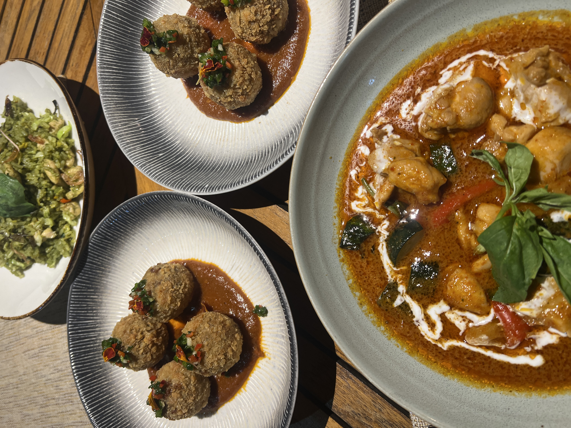 The image shows a wooden table with several plates of food, likely taken in daylight. The plates contain an assortment of dishes. On the left is a plate of green rice and a plate of fried balls with a sauce and garnishes. Next to them is another plate of fried balls in sauce. On the right is a plate of chicken and vegetable curry with basil garnish and a drizzle of coconut milk. The precise location in North Kuta, Indonesia is not discernable from the image. No people, actions, or interactions are shown.