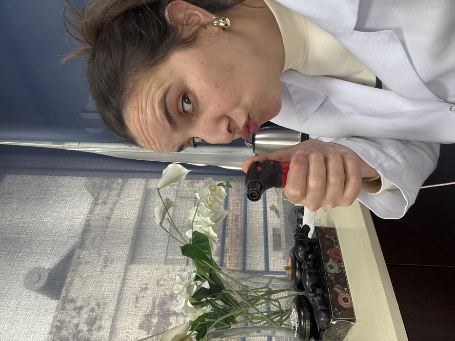 This image captures a female subject in an indoor environment in Meknes, Morocco. The woman, positioned on the right side of the frame, has dark hair pulled back and is wearing a white lab coat over a light-colored turtleneck top, with a gold-colored hoop earring visible on her right ear. Her right eye is wide open and directed at the camera, while her lips are pursed in a pouting expression. She holds a red and black mini torch lighter in her right hand, pointed towards the viewer.
On the left side of the frame, a light-colored countertop displays several objects: a clear glass vase containing white artificial flowers (likely calla lilies or similar), a dark-colored ashtray containing a cigarette butt, and a decorative box with a patterned side, topped by three dark-colored hippopotamus figurines arranged in a line. The background features a window covered by a translucent grey roller blind, through which faint outlines of exterior architecture and a prominent circular shadow are visible. To the right of the blind, a dark blue wall or partition is present, and a stainless steel electric kettle is partially visible behind the woman's right arm.