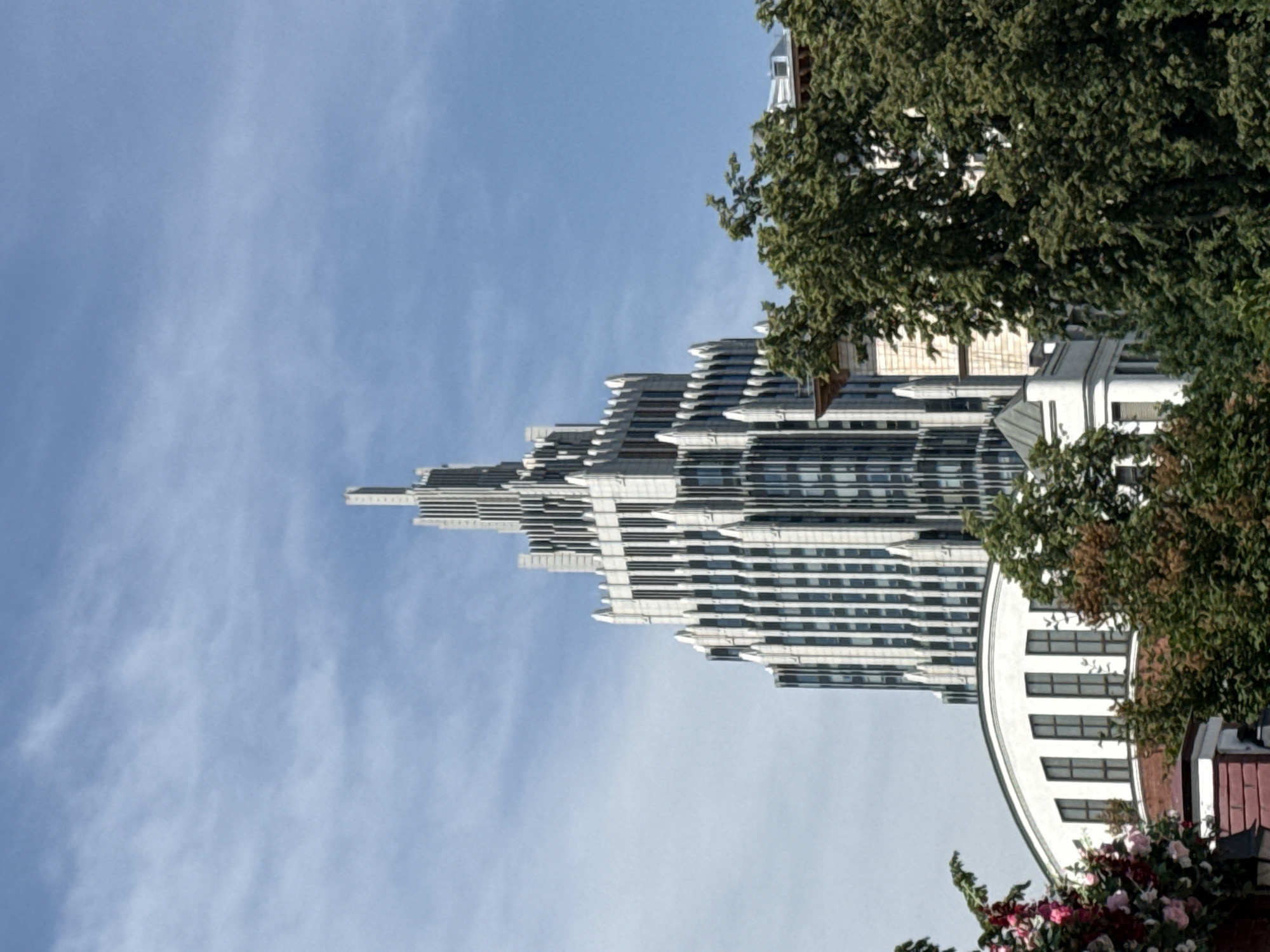An upward-angled, daytime photograph captures a prominent modern skyscraper in Moscow, Russia, framed by lush green foliage and a partly cloudy sky. The central subject is the House on Mosfilmovskaya, a multi-tiered skyscraper characterized by its Art Deco-inspired design, featuring light-colored cladding, numerous vertical window bands, and a distinctive stepped profile culminating in a pointed spire. The building occupies the upper-middle and upper-right portions of the frame, with its lower sections obscured.
In the midground and foreground, a dense canopy of deciduous trees, predominantly green with some autumnal reddish-brown hues on the left, conceals the base of the skyscraper and other structures. To the lower-left, partially visible through the trees, is a white building with a distinctive curved roofline and several vertical rectangular windows, resting on a reddish-brown brick base. To the lower-right, behind the trees, parts of other less defined buildings or structures are discernible. The sky above is a clear blue, interspersed with wispy white clouds that suggest fair weather. No people or animals are visible in the scene.