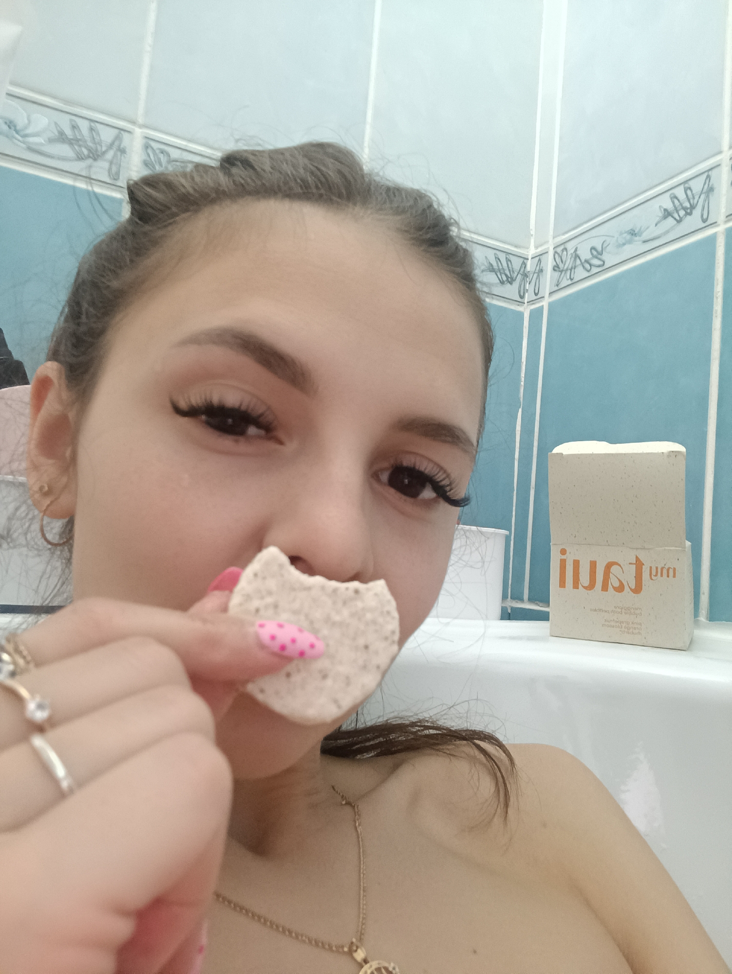 The image is a close-up, seemingly a selfie, taken indoors in a bathroom. A fair-skinned young woman is the central figure, holding a bite-sized piece of what appears to be a rough, light-colored cookie or cracker in front of her mouth. She has long eyelashes, and her left hand is adorned with rings, with one fingernail painted pink with red polka dots. She is wearing a gold chain around her neck. Behind her, a stack of two boxes is visible on the edge of a white bathtub. The boxes have a white and beige color scheme with the word "JUI" prominently displayed in orange letters on one side, and other smaller text. The bathroom features light blue tiled walls with a decorative border near the top. The location is not specified, and the city is listed as Unknown, Unknown.