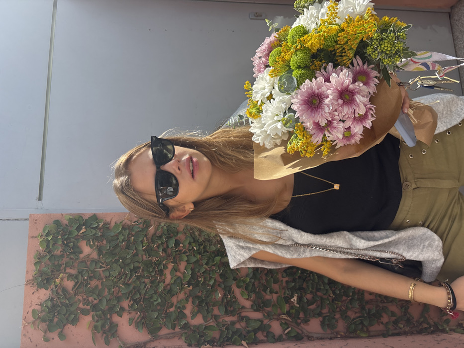 A young adult female is depicted standing outdoors in what appears to be a brightly lit urban or suburban environment, within the city of Aldoar, Foz do Douro e Nevogilde, Portugal. She has long, light brown hair and is wearing black Wayfarer-style sunglasses, a black ribbed tank top, a light grey sleeveless knit vest, and olive green trousers featuring decorative metal eyelets along the sides. A gold chain necklace with a small, light-colored cubic pendant hangs around her neck, and her left wrist displays multiple bracelets, including a wide gold-colored cuff and beaded bands. She is holding a large, mixed bouquet of white, pink, yellow, and green flowers, primarily chrysanthemums, wrapped in brown kraft paper. A set of keys with a colorful patterned ribbon is attached to the flower wrapping. The background to her left features a textured, reddish-pink wall densely covered in dark green ivy, while the background to her right consists of a plain, light grey wall or metal panel.