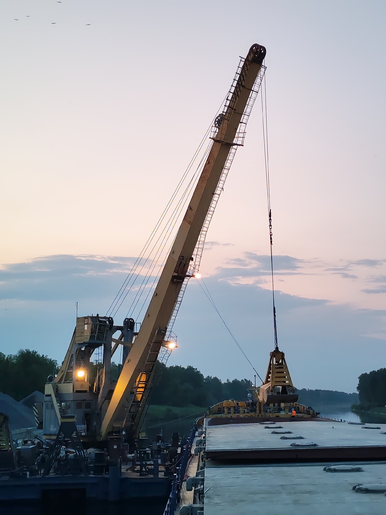A large, yellow crane is positioned on a barge on the Yakhroma canal in Yakhroma, Russia. The crane is equipped with a clamshell bucket suspended from its boom by cables and chains. The bucket appears to be positioned over another barge. A person wearing an orange vest can be seen near the bucket. The background shows trees along the canal bank and a sky with clouds. Several birds are flying in the distance. The crane's lights are illuminated. A pile of material is visible on the barge with the crane.