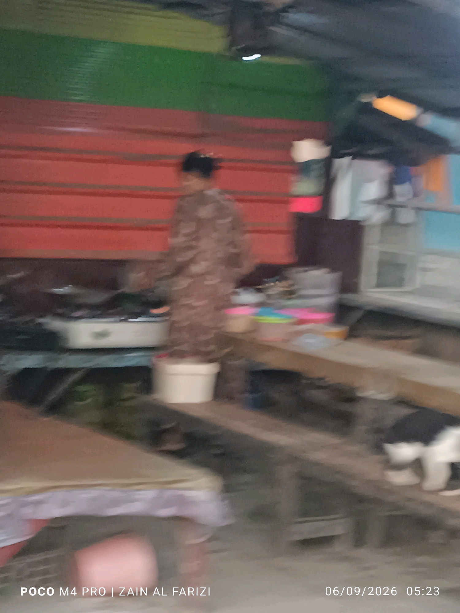 The image depicts an indoor scene, likely a kitchen or food preparation area, with a blurred subject in the background wearing a patterned garment. A red corrugated metal wall is visible behind the subject, with a green band above it. In the foreground, there is a wooden counter or table with various objects, including containers and what appears to be a gas stove. To the right, a partially visible animal, possibly a cat, is present. The overall environment suggests a humble or informal setting. The text overlay at the bottom indicates the image was taken with a POCO M4 PRO by ZAIN AL FARIZI on 06/09/2026 at 05:23. The location is identified as Arosbaya, Indonesia.