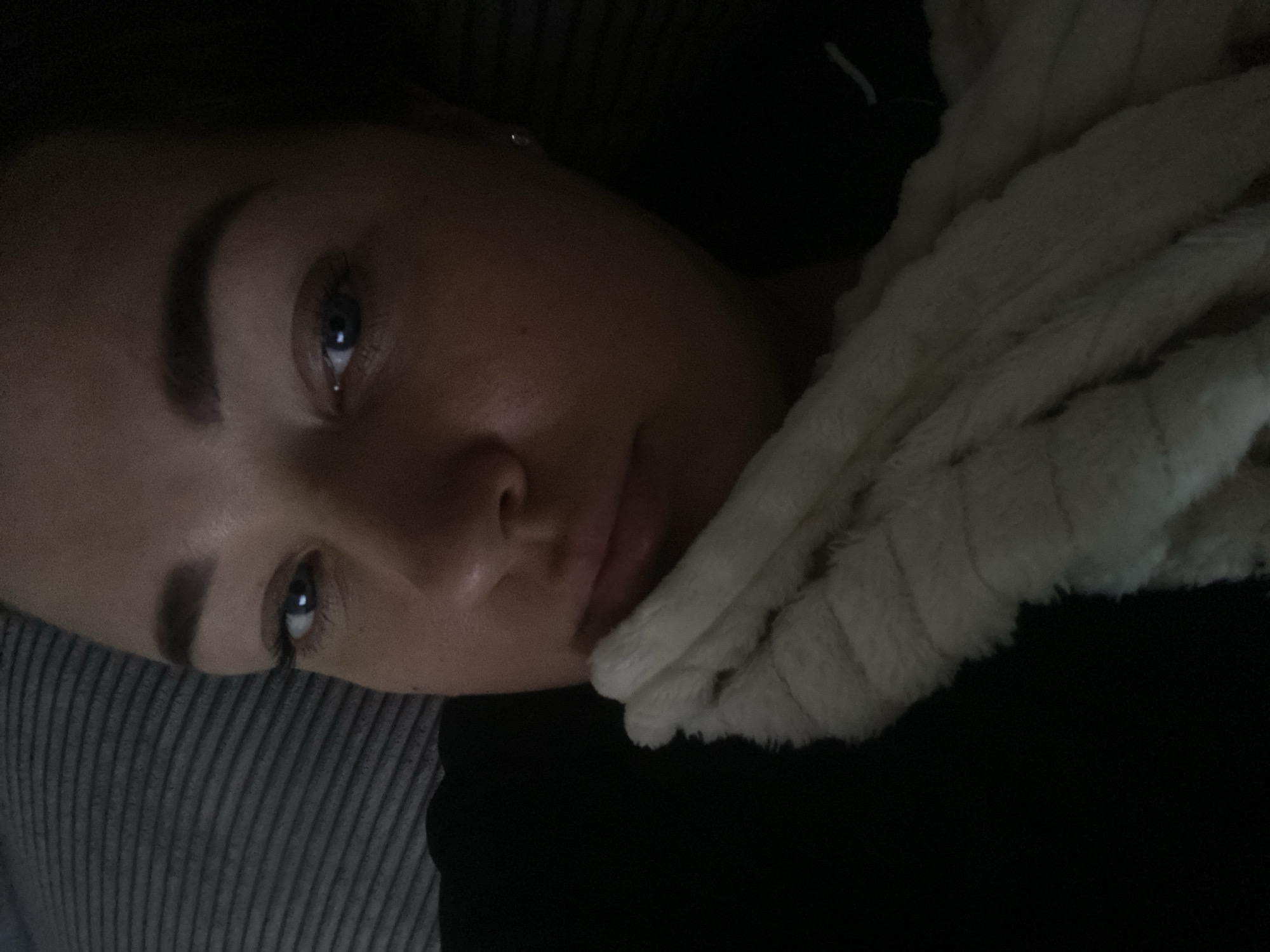 A young woman is lying on her side, possibly on a bed or couch covered in gray striped material. She is resting against a fluffy white blanket or scarf. She is wearing a black shirt. The lighting is dim, focusing mainly on her face. The image does not provide enough details to determine her location in Sochi, Russia.