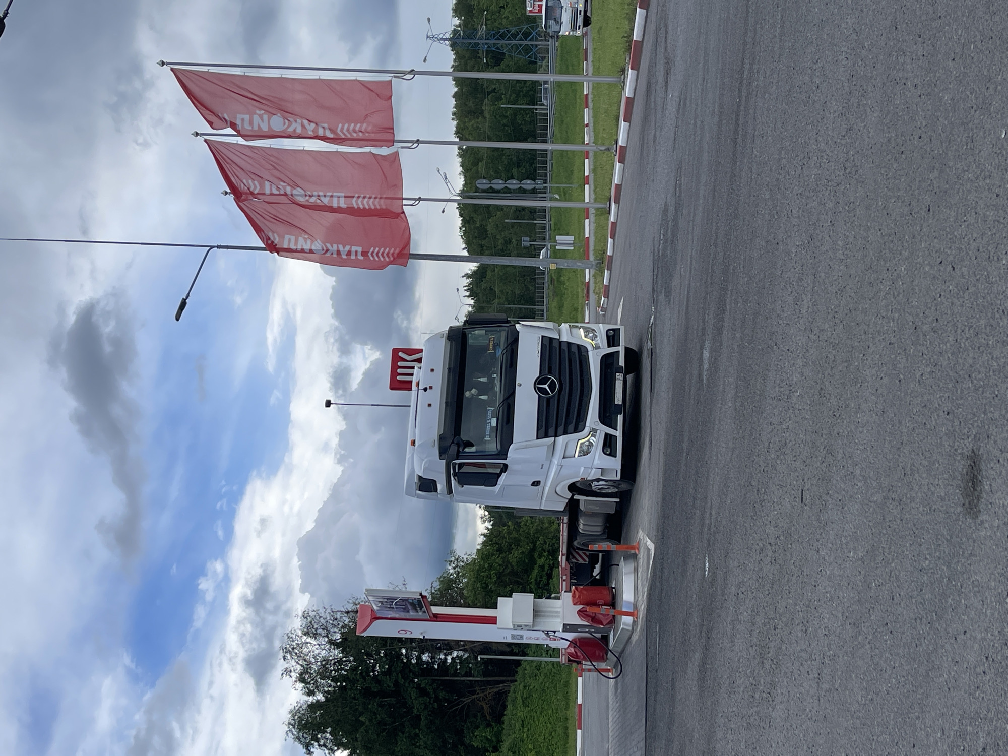 A Mercedes-Benz Actros truck is parked at a Lukoil gas station in Бавыкино, Russia. The truck is white and has a Lukoil logo on top of the cab. There are three red Lukoil flags flying near the gas pumps. The sky is cloudy. A red and white striped curb is adjacent to the gas station. The ground is asphalt. There are green trees and shrubs in the background.