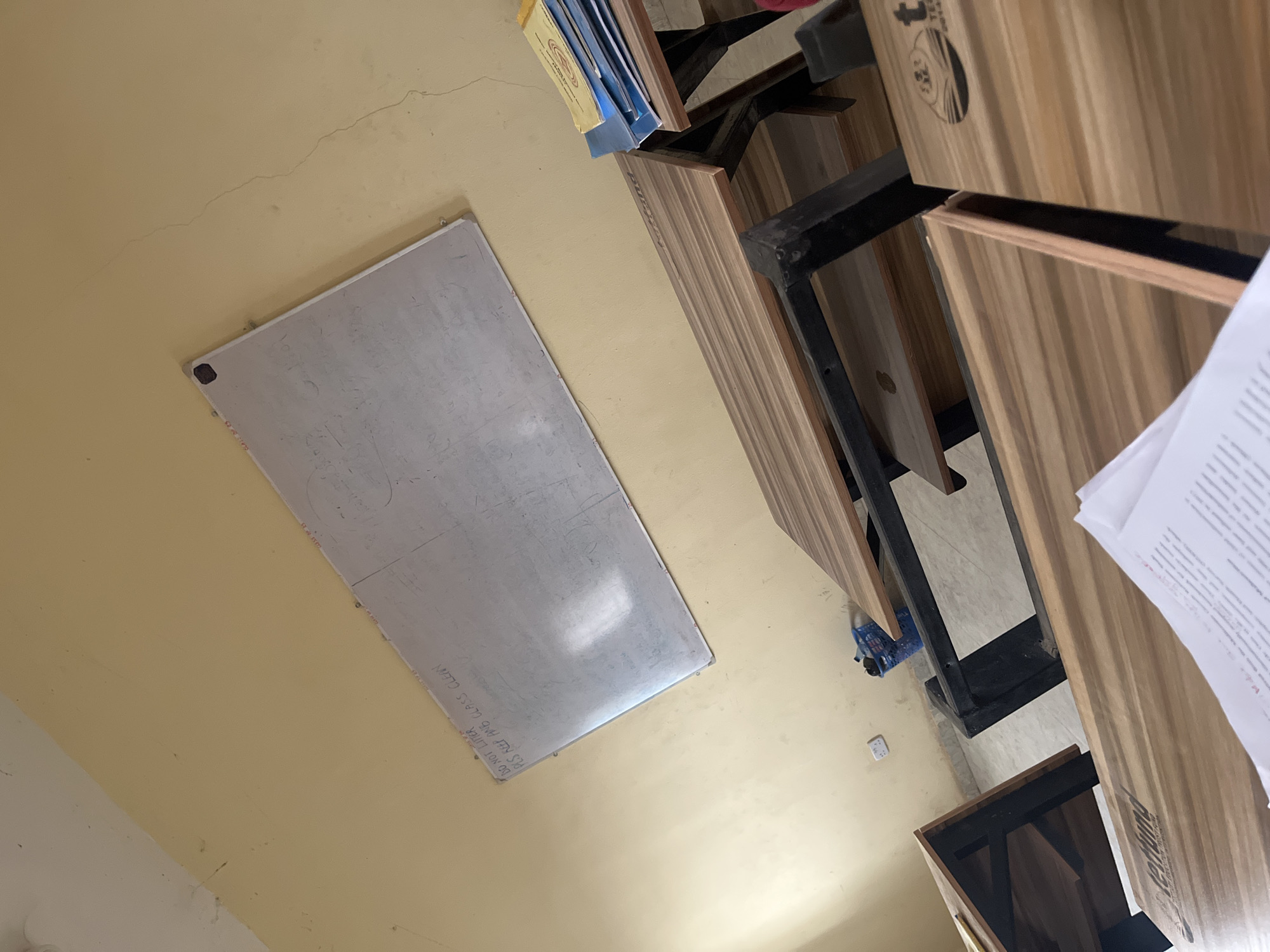 The image depicts an indoor scene, possibly a classroom or office, in the city of Baganakwo, Nigeria. The walls are painted a pale yellow color. A whiteboard is mounted on the wall, with some writing faintly visible. Several desks are arranged in a row, with black metal frames and light brown wooden tops. A stack of blue books is visible on one of the desks. A sheet of paper with printed text is partially visible on the right side of the frame. There are no people visible in the scene.