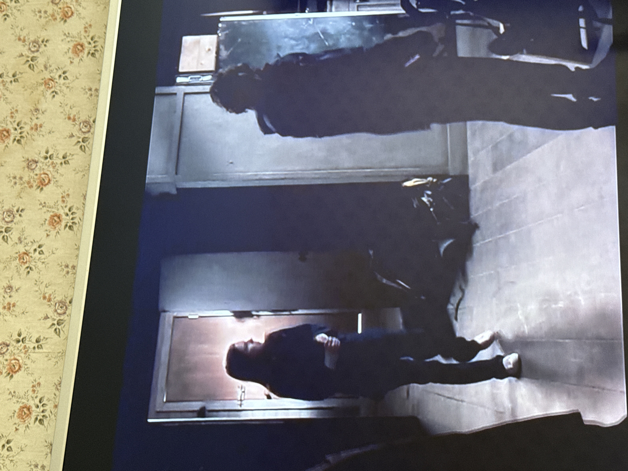 A photograph shows two figures lying down in a dimly lit interior space. One figure is near a door, and the other figure is near a cabinet or table. The figures appear to be lying on the floor. The environment is not definitively identifiable, and location information about Mytishchi, Russia, cannot be inferred from the visual data.