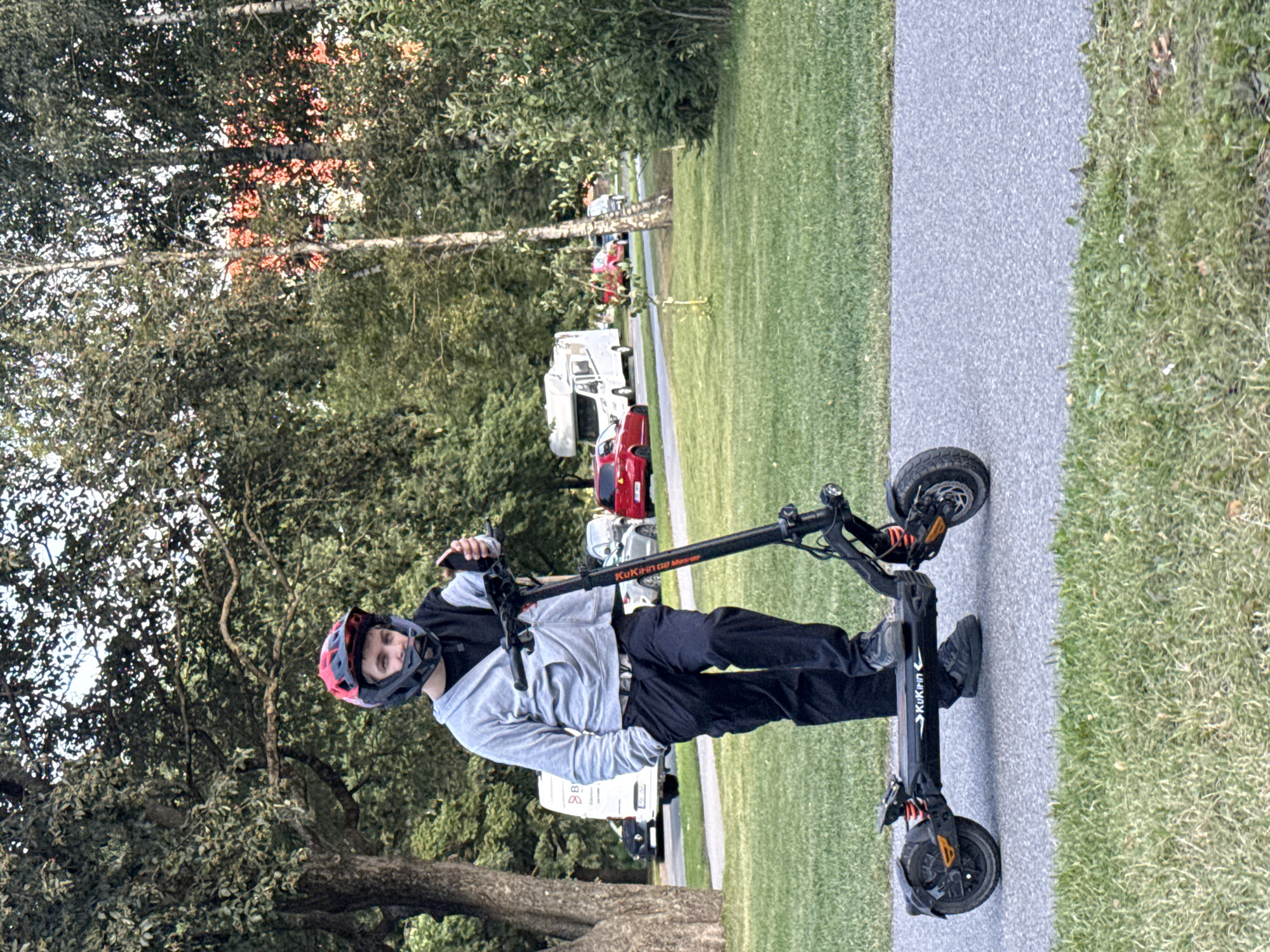A young person wearing a red and black helmet and a gray hooded sweatshirt stands on an electric scooter on a paved path. The person is holding a smartphone in their right hand. The scooter is black with orange accents and the brand name "Kukirin" is visible. In the background, there is a grassy area with trees, and several vehicles are parked or driving on a road, including a white camper van, a red car, and a black car. The setting appears to be a park or a recreational area within a city.