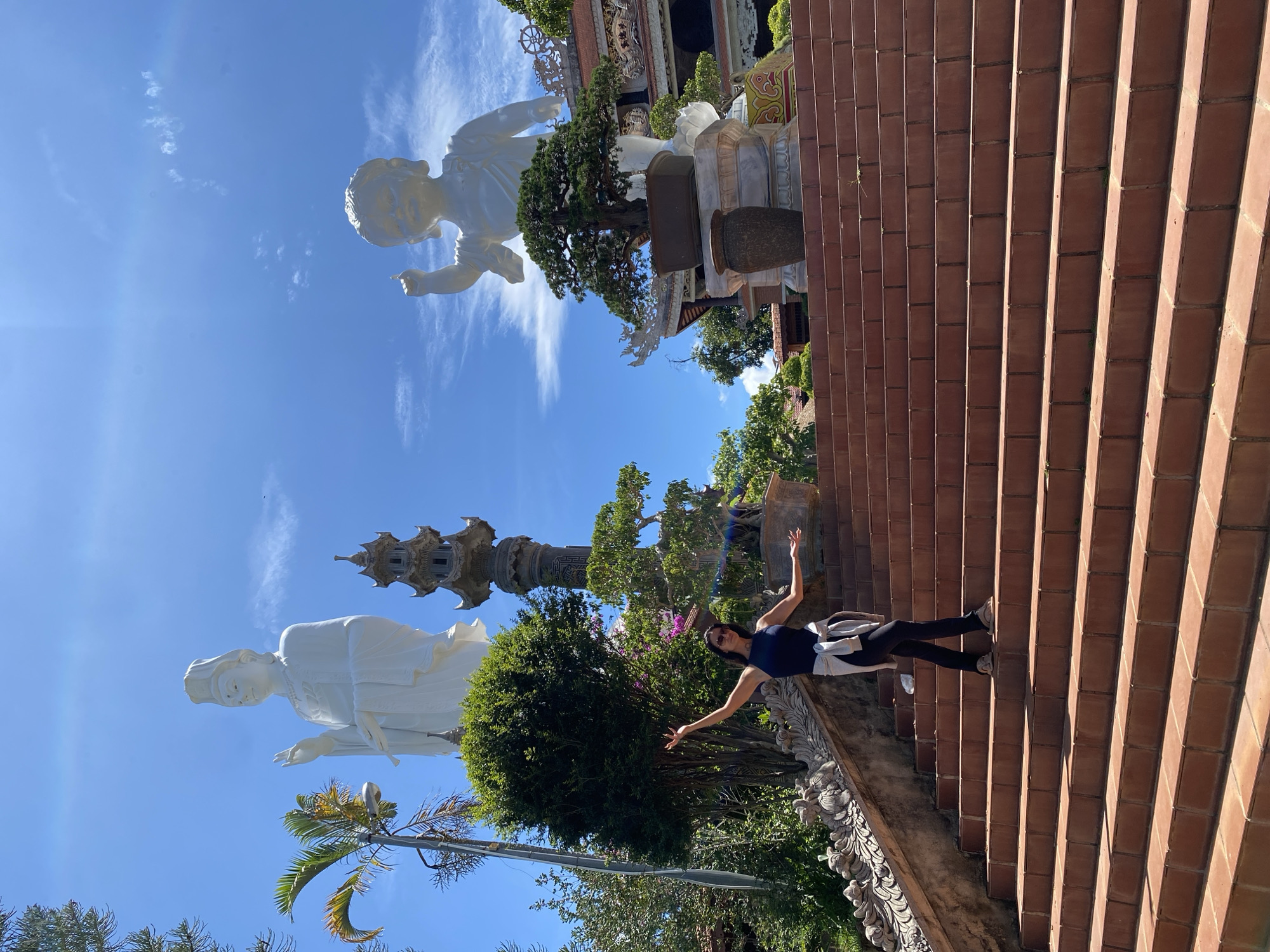 The image shows a young woman standing on a set of terracotta stairs with her arms outstretched. She is wearing a dark blue tank top, black leggings, and white sneakers. A light-colored jacket is draped over her shoulders. She is looking towards the camera and appears to be smiling, conveying a sense of joy and freedom.
In the background, two large white statues are visible against a clear blue sky with scattered white clouds. The statue on the left is a seated figure with a serene expression, wearing traditional Asian attire and a headdress. The statue on the right appears to be a smaller, childlike figure in a dynamic pose. A tall, multi-tiered pagoda-like structure is also visible between the statues. Lush green trees and flowering bushes surround the statues, adding to the vibrant atmosphere.
The scene is set outdoors, likely during the daytime given the bright sunlight and clear sky. The terracotta stairs suggest a temple or religious site. The overall impression is one of cultural tourism and enjoyment of a scenic and spiritual location in Thôn 5, Vietnam.