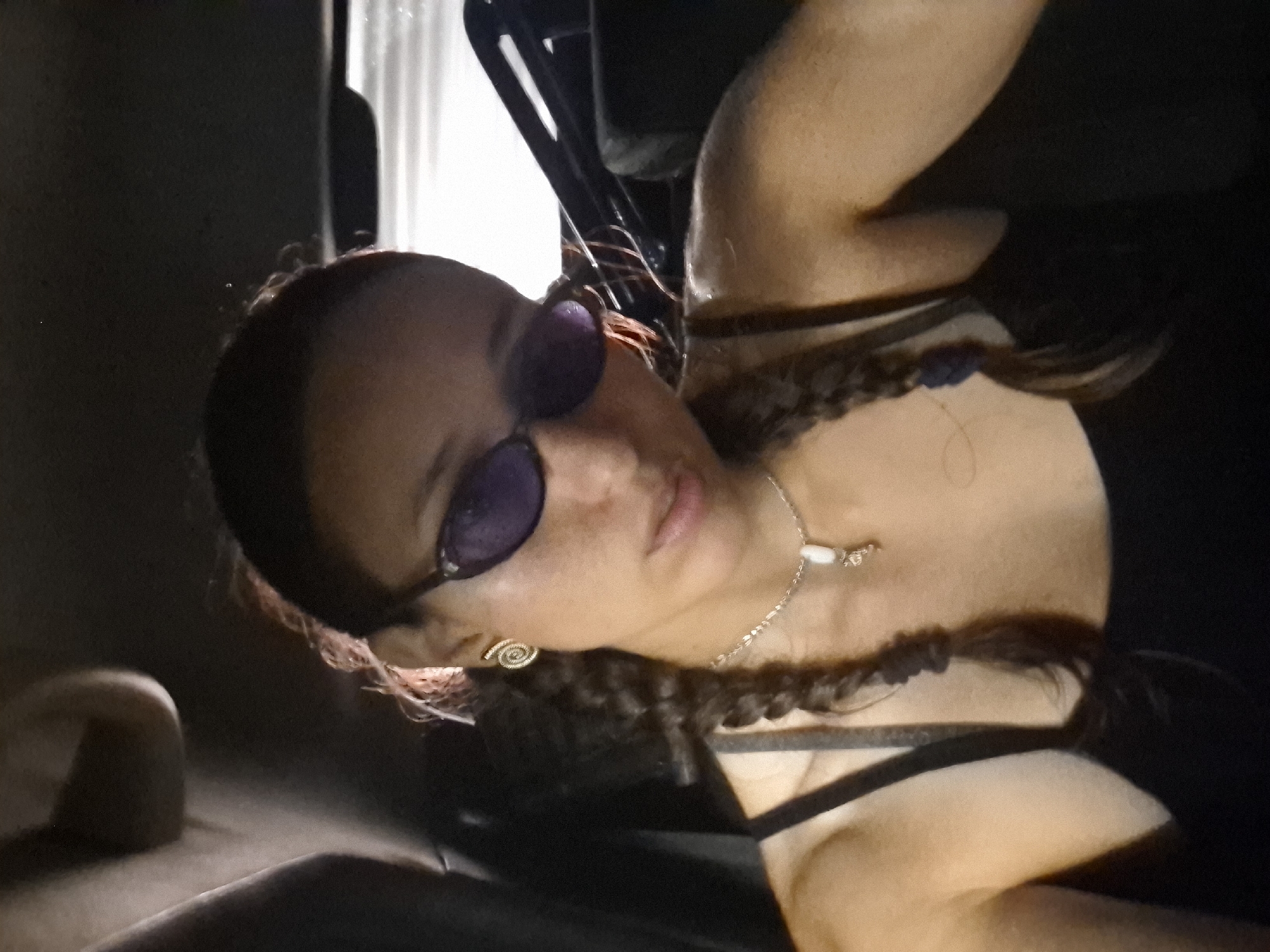A woman with brown hair braided and wearing sunglasses is sitting indoors in a vehicle. She has a silver chain necklace with a light-colored charm. She is wearing a black top with thin straps. A small, gold-colored, spiral-shaped earring is visible in her left ear. The location is unknown, and not determinable based on the image.