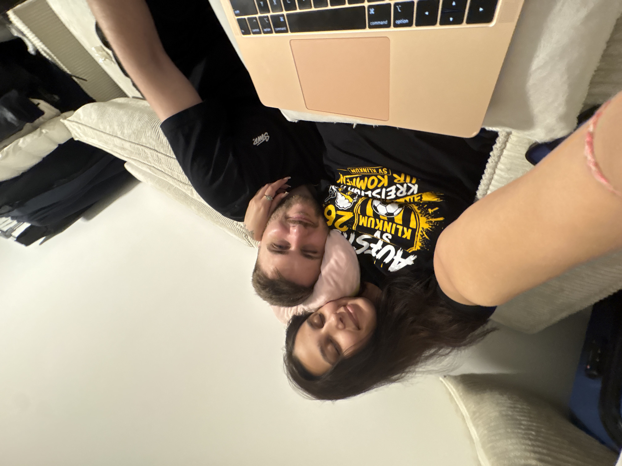 This image depicts a couple lounging on a sofa, with a laptop positioned in front of them. The woman is lying down, her head resting on a pink pillow, her eyes closed. The man is lying next to her, with his head also on the pillow, looking towards the camera. They are both wearing dark t-shirts. The t-shirt worn by the man has a graphic design with text that appears to be in German. The sofa is upholstered in a light-colored, ribbed fabric. In the background, on the left, there are clothes hanging on what appears to be a closet rod. The scene is indoors, likely in a residential setting. The presence of the German text on the t-shirt strongly suggests the location is in Germany. Mönchengladbach is a city in Germany, making it a plausible location for this scene.