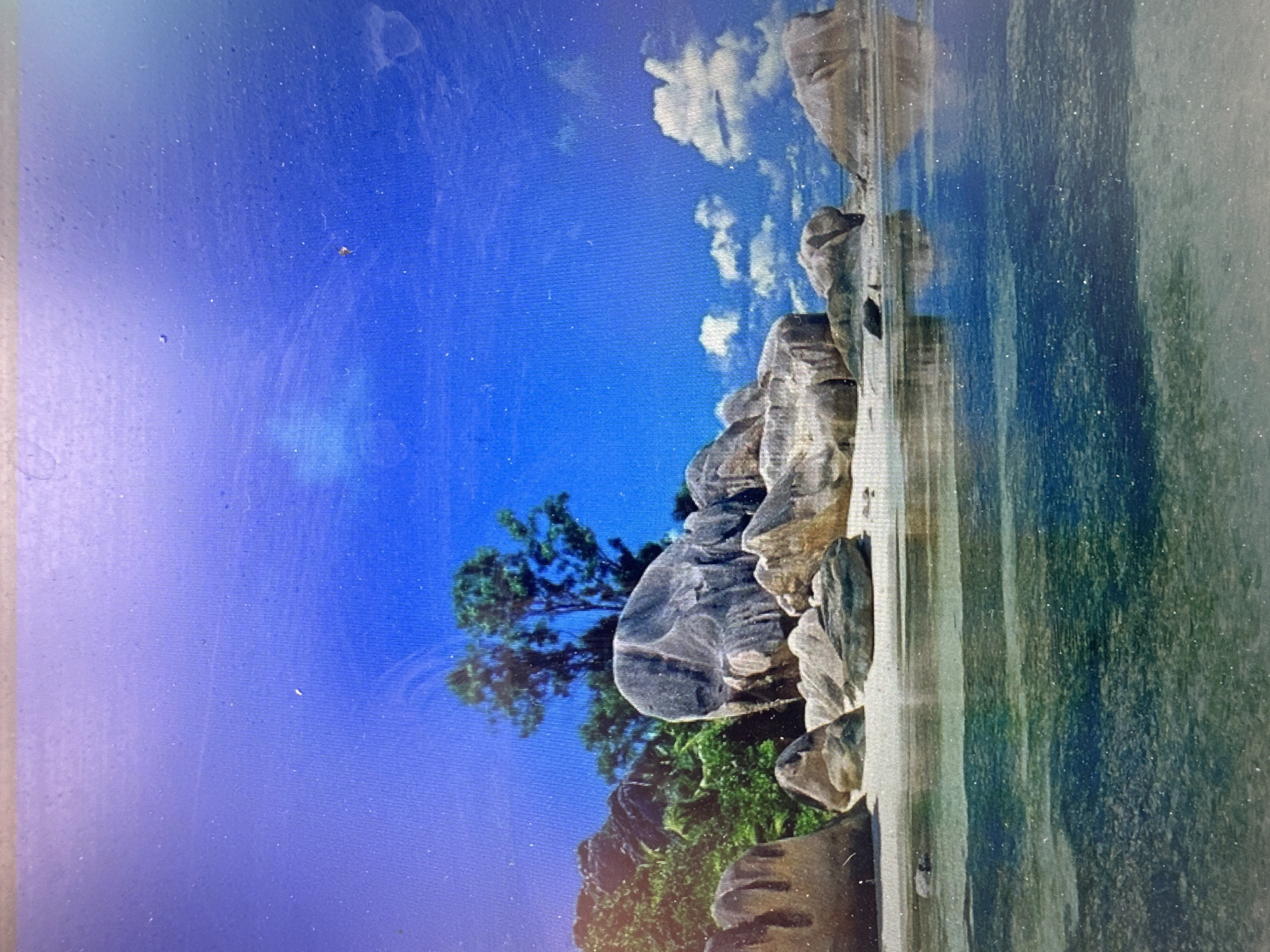 A photograph captures a tropical beach scene under a bright blue sky. Large, weathered granite boulders are scattered across a sandy shore, with lush green foliage and tall trees forming a backdrop. The clear turquoise water gently laps the beach, reflecting the sky and the rocks. The overall impression is one of a serene and idyllic natural landscape. There are no people or man-made structures visible in the image. The location is not Grigorevka, Kyrgyzstan, as this image depicts a tropical beach, which is not characteristic of that region.