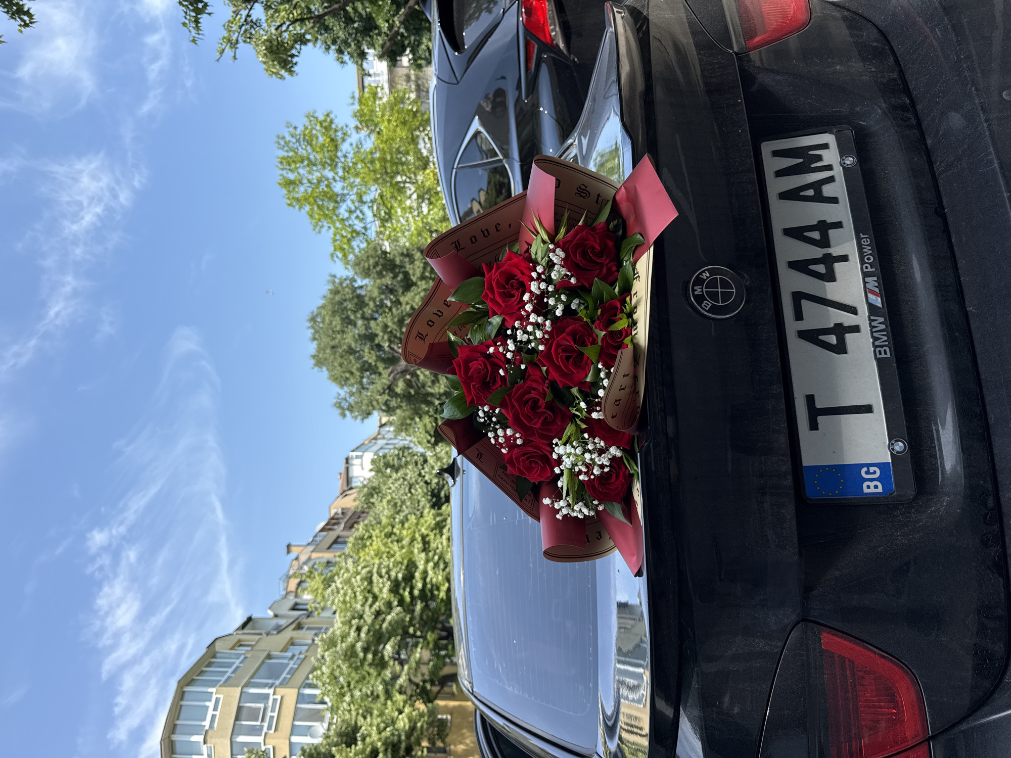 A black BMW car, appearing dusty, is parked outdoors on a bright, sunny day. A vibrant bouquet of red roses and white baby's breath, wrapped in brown paper with a red ribbon, is placed on the trunk of the car. The wrapping paper has partially visible text including "Love," "tart," "St," "you more," and "an say."
The car's license plate, which is from Bulgaria (indicated by "BG" and the EU stars), reads "T 4744 AM". The license plate frame also displays "BMW M Power" and the BMW logo. The car's tail lights are visible on the right side of the trunk.
In the background, under a clear blue sky with some wispy white clouds, there are green trees and parts of multi-story buildings, suggesting an urban or suburban environment consistent with Varna, Bulgaria. The overall scene captures a moment likely involving a gift or special occasion, highlighted by the striking bouquet against the dark car and bright outdoor setting.
