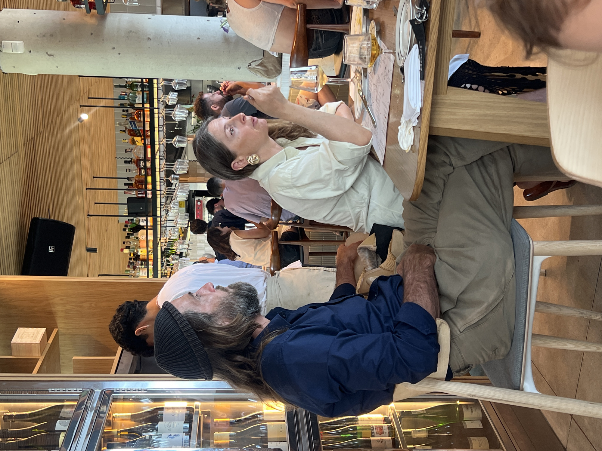 The image captures an indoor scene at a restaurant, likely in Tel Aviv, Israel given the architecture and clientele. A group of people are seated at a wooden table, seemingly engaged in a meal. The woman in the white blouse is holding something in her hand while talking. A man with a beard is wearing a blue shirt and a beanie seated near the front of the image next to a wine cooler cabinet. In the background, shelves are filled with various bottles, likely alcoholic beverages. A black speaker is visible on the wall. The environment suggests a relaxed, social atmosphere, possibly during lunchtime.