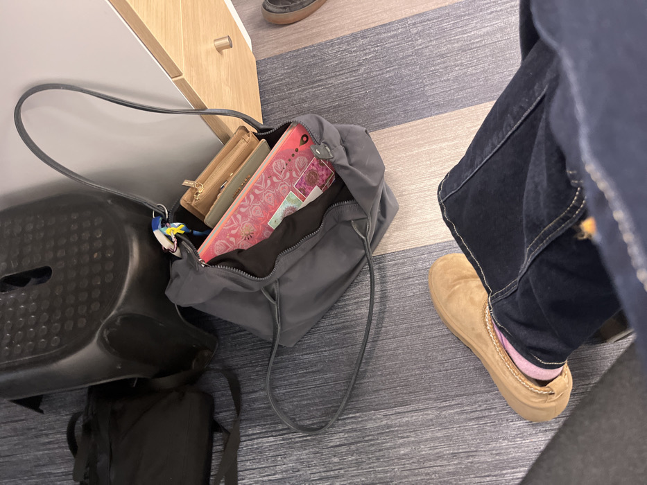 A gray nylon tote bag sits on a striped floor, next to a small black plastic step stool and a closed cabinet. A wallet, book, and other items are visible in the open bag. A black backpack is partially visible under the stool. A person wearing dark wash jeans and tan slip-on shoes with pink socks stands next to the bag. The floor is light gray with blue and tan stripes.