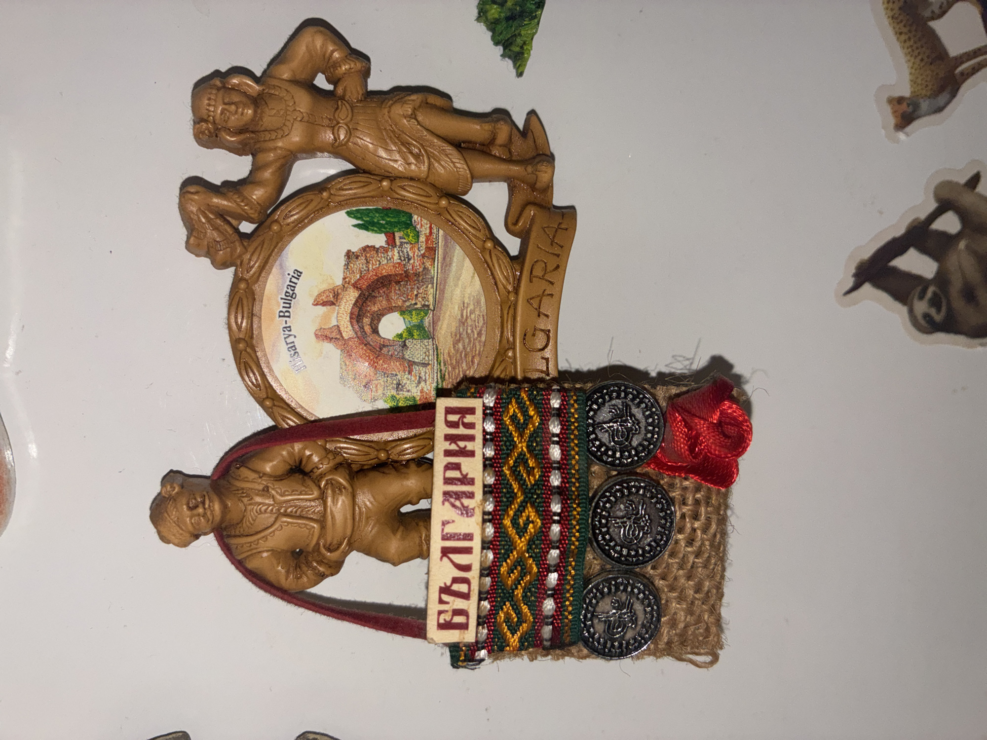 An ornate souvenir magnet is adhered to a white surface. The magnet features two figures, a male and a female, both sculpted in a brown material resembling wood or clay. The male figure, on the left, wears traditional attire and holds a small, burlap-like bag adorned with colorful embroidered fabric and three silver-colored coins. A red ribbon hangs from the bag. The female figure, on the right, is in a dancing pose, holding a scarf aloft. Behind them, a circular frame depicts a scene of ancient ruins with the text "Hisarya - Bulgaria" and "BULGARIA" across the bottom. The background of the white surface has a few scattered stickers of animals, including a sloth and a cheetah, visible at the bottom. The location of the souvenir is not specified but it features Bulgarian imagery.