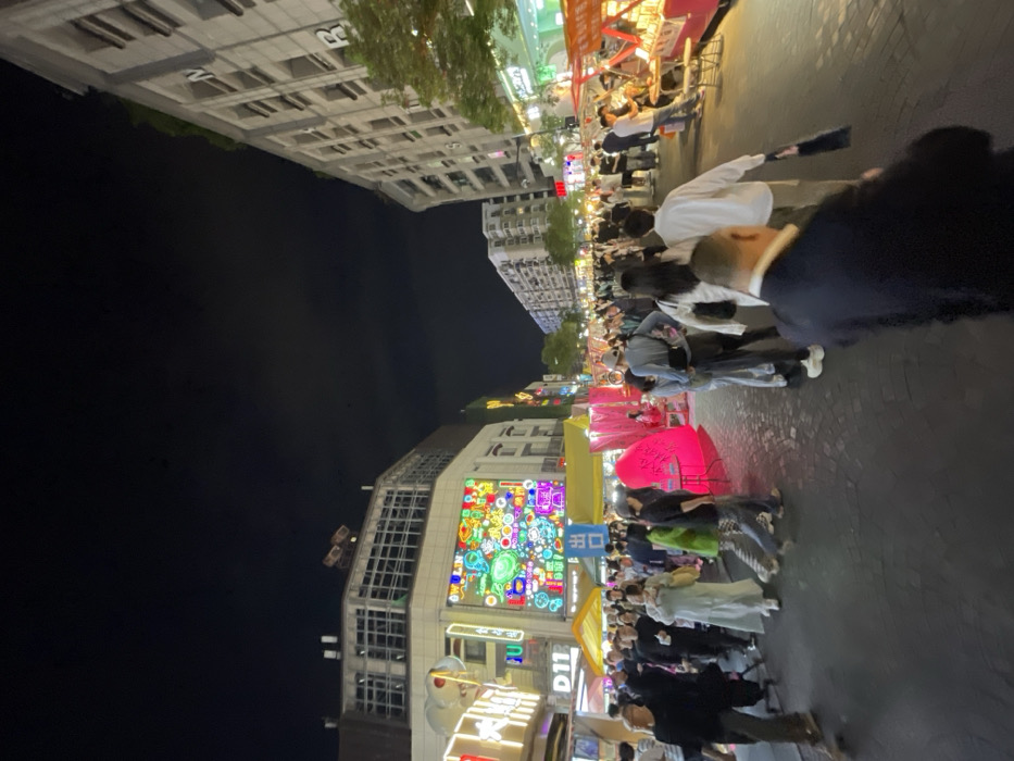 The scene is an evening night market on a street in Hangzhou Shi, China (30.264, 120.156). The market is crowded with many people walking around, shopping, and eating at food stalls. Some people are carrying bags, others are eating snacks, and several have umbrellas. A variety of food stands line the sides of the street, and some have brightly lit signage. The street has a cobblestone surface and the buildings are tall and lined with many windows. There is a brightly lit, multi-colored sign on the side of the building at the left of the scene. The sign has many squares, with some of them lit in bright neon colors. The sky is dark and the night is clear.