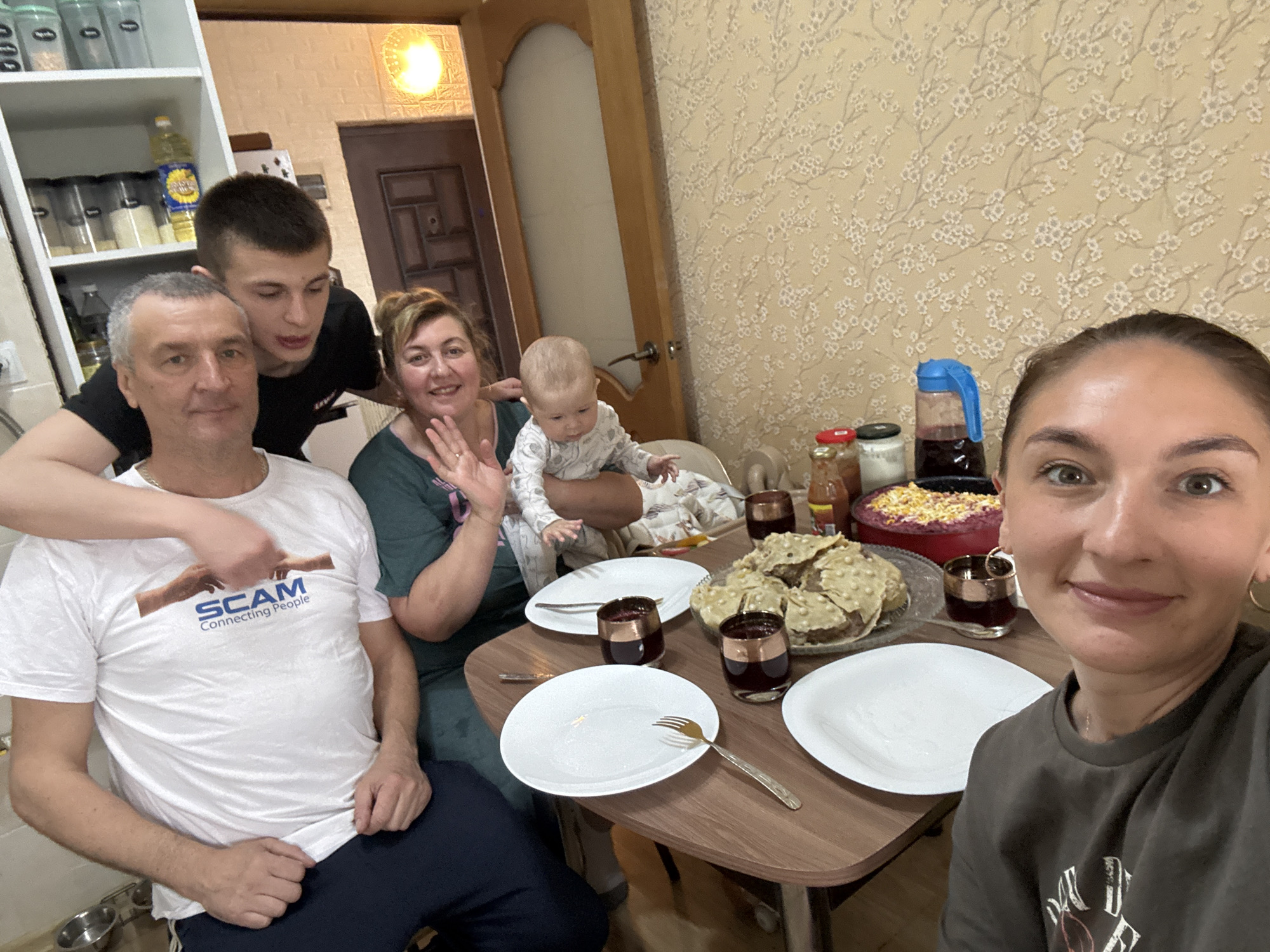 The image shows a group of six people gathered around a table in what appears to be a home setting. The environment includes a white cabinet, patterned wallpaper, and a doorway leading to another room.
From left to right:
1. An older man sits at the table, wearing a white T-shirt with a design that says "SCAM Connecting People."
2. A younger man stands behind him, with his arm resting on the older man's shoulder.
3. A woman is holding a baby. She is smiling and waving.
4. A baby is sitting on the woman's lap, looking at the camera.
5. A woman is taking the photo, holding a camera.
The table is set with white plates, glasses filled with a dark liquid, and a cake. There is also a red bowl containing food. The photograph appears to be taken at someone's home; the location is Sochi, Russia.
