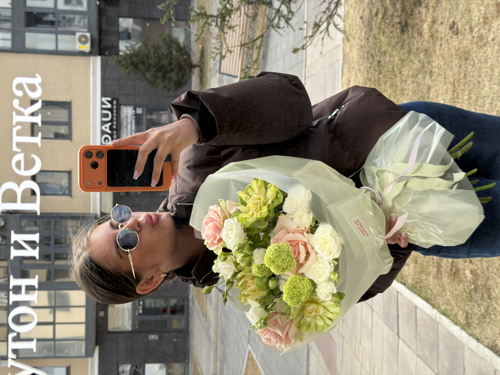 A woman in sunglasses and a brown jacket is holding a bouquet of roses, light green dahlias, and pompom chrysanthemums. She is taking a selfie with a phone in an orange case. The bouquet is wrapped in translucent white paper. The background includes a building with text in Russian, likely a business name, and a grassy area with a sidewalk. This is Krasnoyarsk, Russia.
