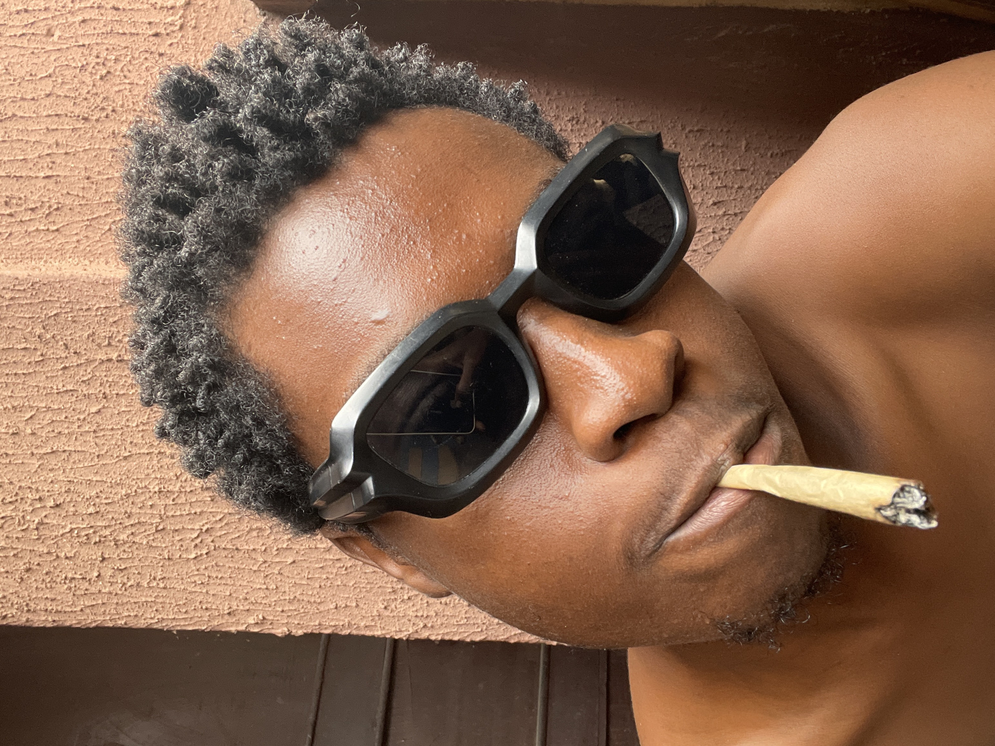 This media file features a close-up, high-angle shot of a Black person, presumably male, lying down and smoking a hand-rolled cigarette or joint. The person is wearing black, angular sunglasses and has short, curly, dark hair. The skin appears to be tanned and slightly dewy, suggesting warm weather.
The background is textured, appearing to be a terracotta-colored wall with a dark wooden surface below. The lighting suggests it is daytime, possibly late afternoon due to the warm tones and shadows. The setting hints at an outdoor or semi-outdoor environment, possibly a patio or a wall outside a building.
The person's face is turned to the side, with the joint held between their lips. Their expression is neutral, and the overall mood could be interpreted as relaxed or contemplative. No text is visible in the image. The location context provided is Nkpor, Nigeria.