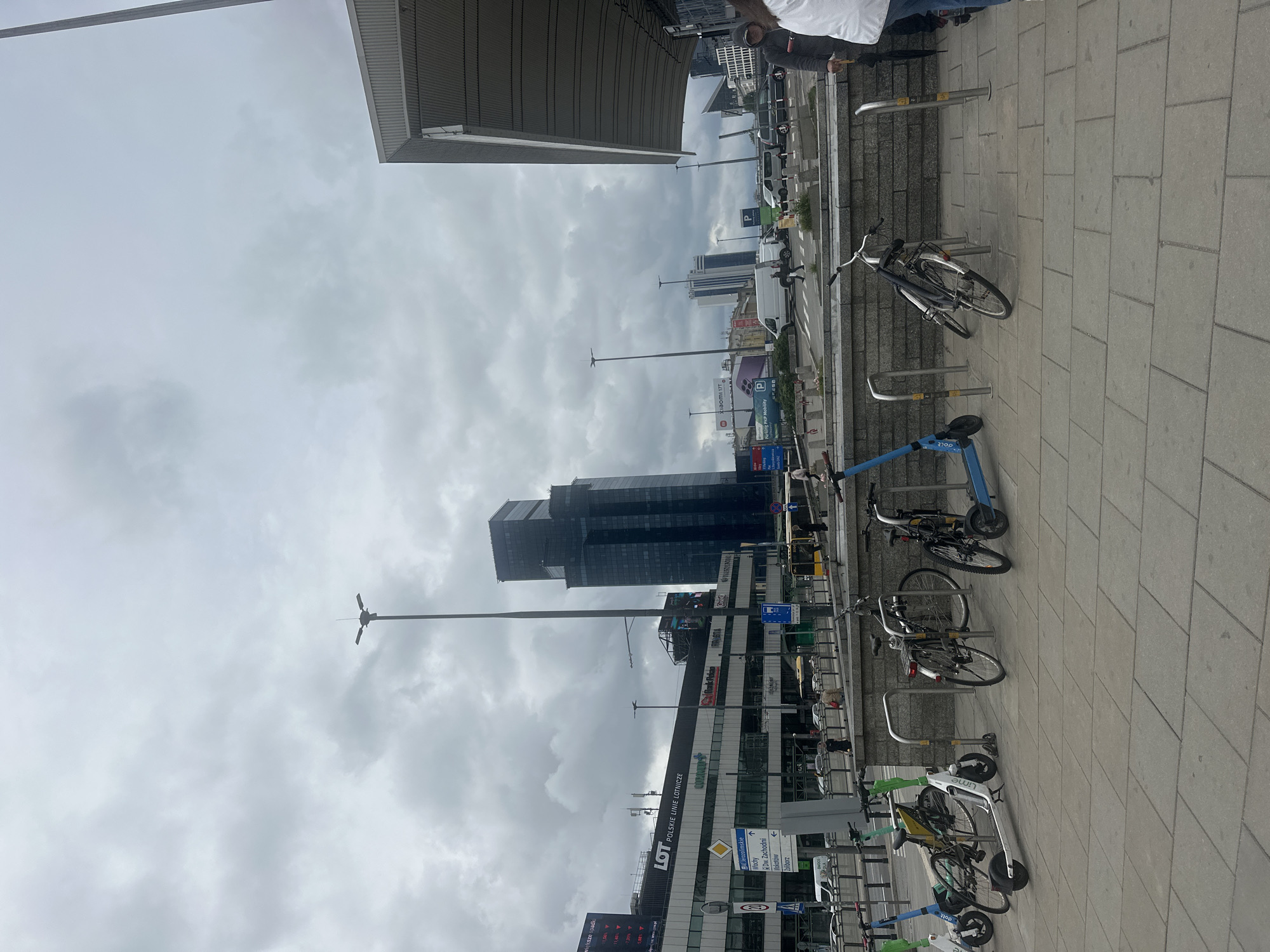 This media file captures a street scene in Warsaw, Poland, likely on an overcast day with a cloudy sky. In the foreground, a row of bicycles and electric scooters are parked at a rack. The objects are varied in color and type, suggesting a public rental service. Beyond the parking area, there's a street with a few cars and a white van, and further in the background, modern high-rise buildings dominate the skyline, one of them a dark, glassy skyscraper. Signs for "LOT POLSKIE LINIE LOTNICZE" (LOT Polish Airlines) are visible on one of the buildings, along with other commercial signage like "XIAOMI 177" and "P" indicating parking. A tall street lamp with multiple lights stands prominently against the sky. The overall impression is one of a busy urban environment, possibly near a transportation hub like a train station, given the context clues and the presence of various modes of transport. The lighting suggests it is daytime, but the cloud cover creates a muted, diffused light. No specific emotions are discernible from the people who are either partially visible or distant figures on the street.