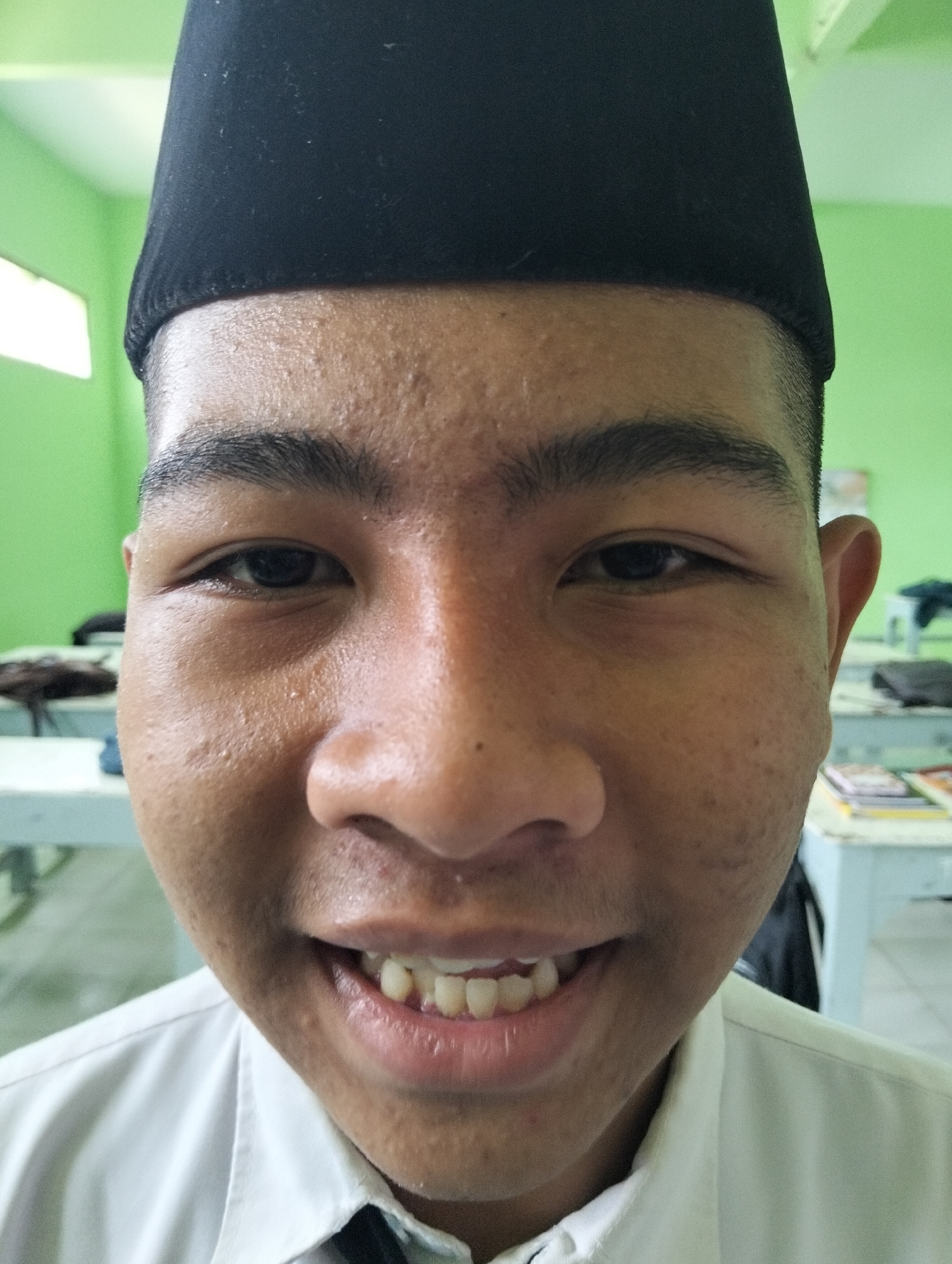 A medium shot, head and shoulders portrait of a young Indonesian male in Kranji, Indonesia, is captured from a slightly low angle. The subject is wearing a black peci hat and a white collared shirt. He is looking directly at the camera and smiling broadly, revealing his teeth. His face is close to the lens, filling most of the frame. The background is out of focus, showing a light green wall and what appears to be a classroom setting with tables and chairs. The lighting is bright, with some skin shine visible on the subject's forehead and cheeks.