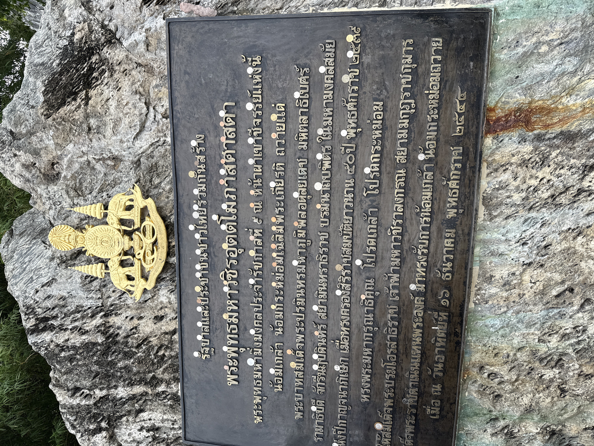 The image displays a commemorative plaque affixed to a large, rough-textured rock face. Above the plaque, a golden emblem, likely a royal crest, is mounted on the rock. The plaque itself is dark, inscribed with Thai script in a lighter color, and features numerous small coins embedded around the text. The rock face is greyish-white with dark grey and black veins, and some green staining is visible on the lower portion. In the background, dense green foliage, possibly bamboo, is partially visible at the top left. The overall scene suggests an outdoor monument or memorial. The plaque is located in Na Chom Thian, Thailand.