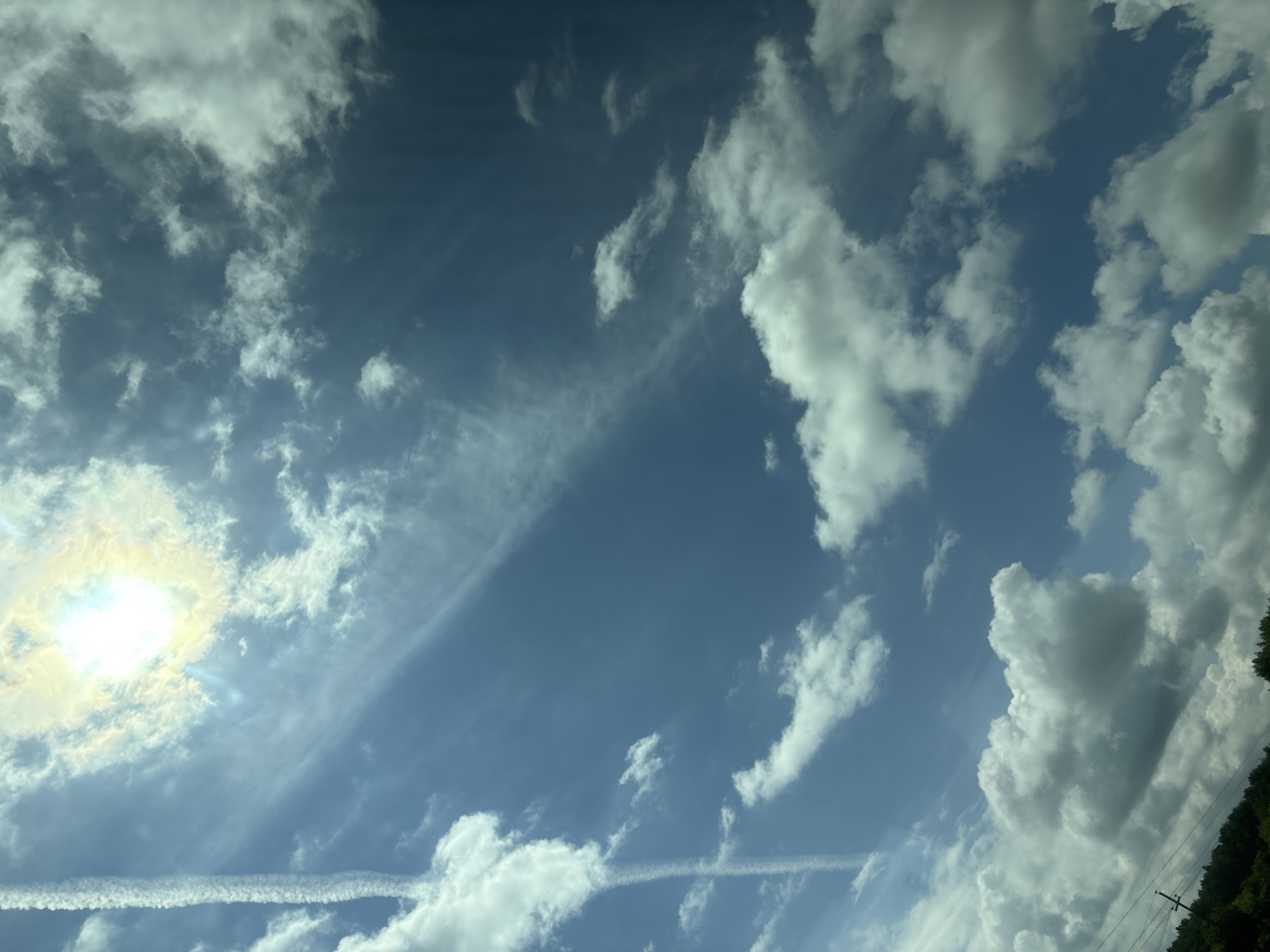 The image shows a bright sun with a halo effect in the upper left quadrant, set against a vast blue sky filled with scattered white and grey clouds. A prominent contrail from an aircraft cuts vertically through the left side of the sky. The lower portion of the image reveals a sliver of lush green trees and vegetation, with visible power lines and utility poles in the far lower left corner. The scene depicts an outdoor environment under a partly cloudy sky, possibly a rural or suburban area, given the presence of trees and utility infrastructure. The specific location in Таймурзино, Russia, is not visually discernible from the provided image alone.
