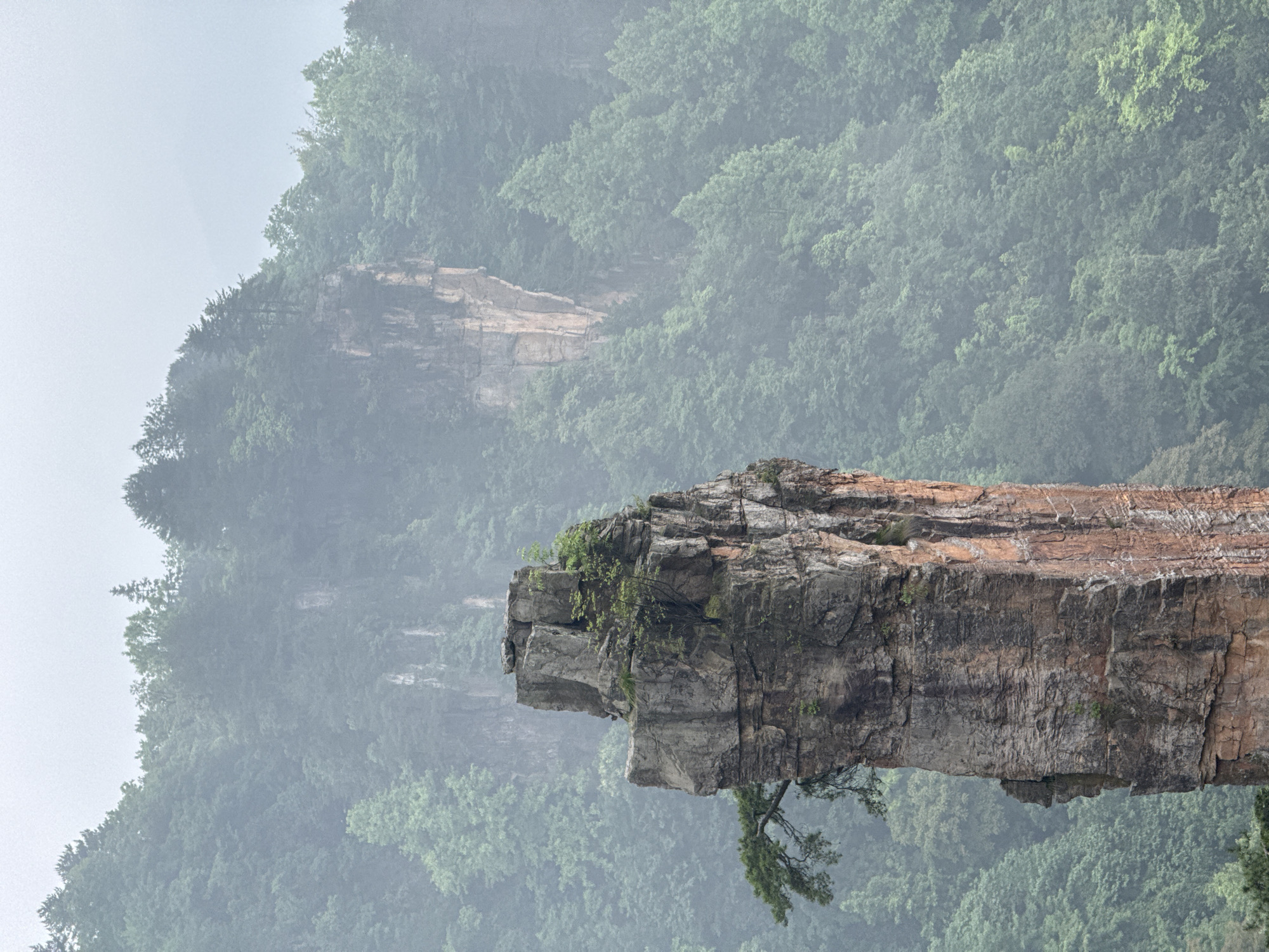 The image depicts a dramatic natural rock formation, specifically a narrow, overhanging cliff, possibly part of a pillar or promontory. The scene is set in a mountainous region with dense, green vegetation, suggesting a forested landscape. The atmosphere appears misty or foggy, obscuring the background mountains. The cliff face has multiple layers and textures, including some vegetation.