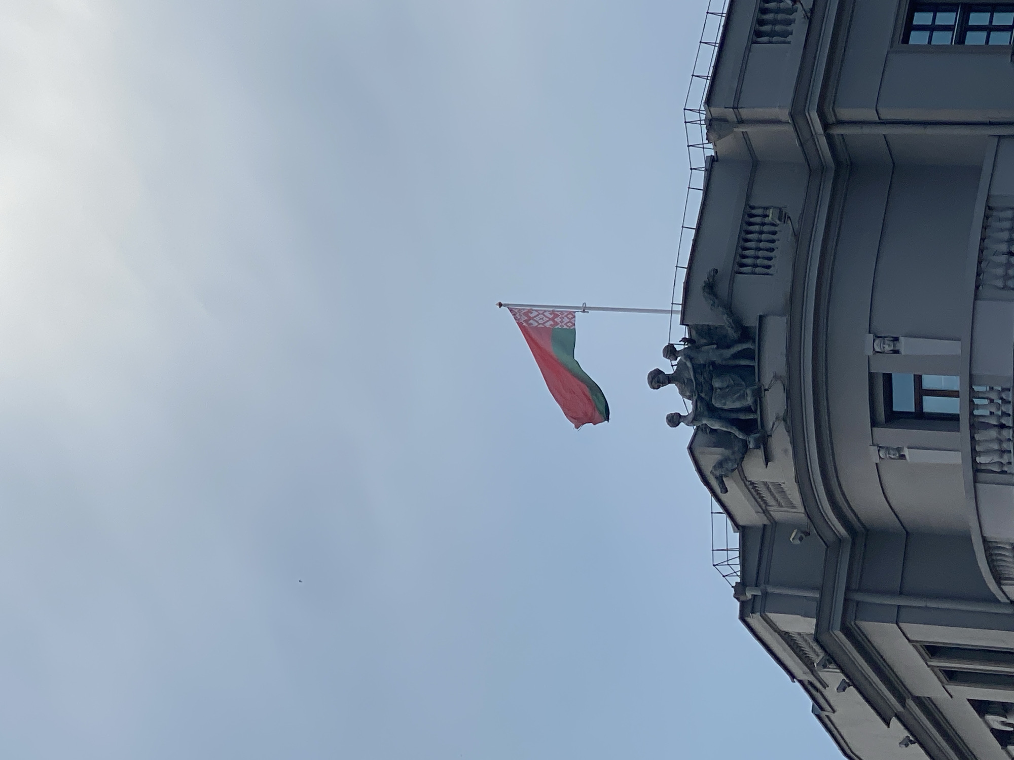 The image captures a view looking upwards at the facade of a grand building with a clear sky in the background. A flag, appearing to be the national flag of Belarus with its red and green stripes and white ornamental pattern, is prominently displayed, attached to a flagpole. The flag is in motion, suggesting a gentle breeze.
The building features classical architectural elements, including curved sections, decorative cornices, and rows of windows. On a prominent ledge of the building, there are several sculpted figures of people. These figures appear to be part of a larger monument or embellishment, possibly depicting historical or symbolic representations. The style of the sculpture and the building suggests an important or historical landmark.
The lighting indicates it is daytime, likely late morning or afternoon, given the brightness of the sky. The sky is a pale blue with a slight haze, suggesting it might be a partly cloudy or overcast day, though no distinct clouds are visible. There are no discernible people actively engaged in any activity in the immediate vicinity of the building's facade as seen in the frame. The overall impression is one of solemnity and grandeur, associated with national symbols and historical architecture. The location is confirmed as Minsk, Belarus, which aligns with the presence of the Belarusian flag and the architectural style often found in the city.