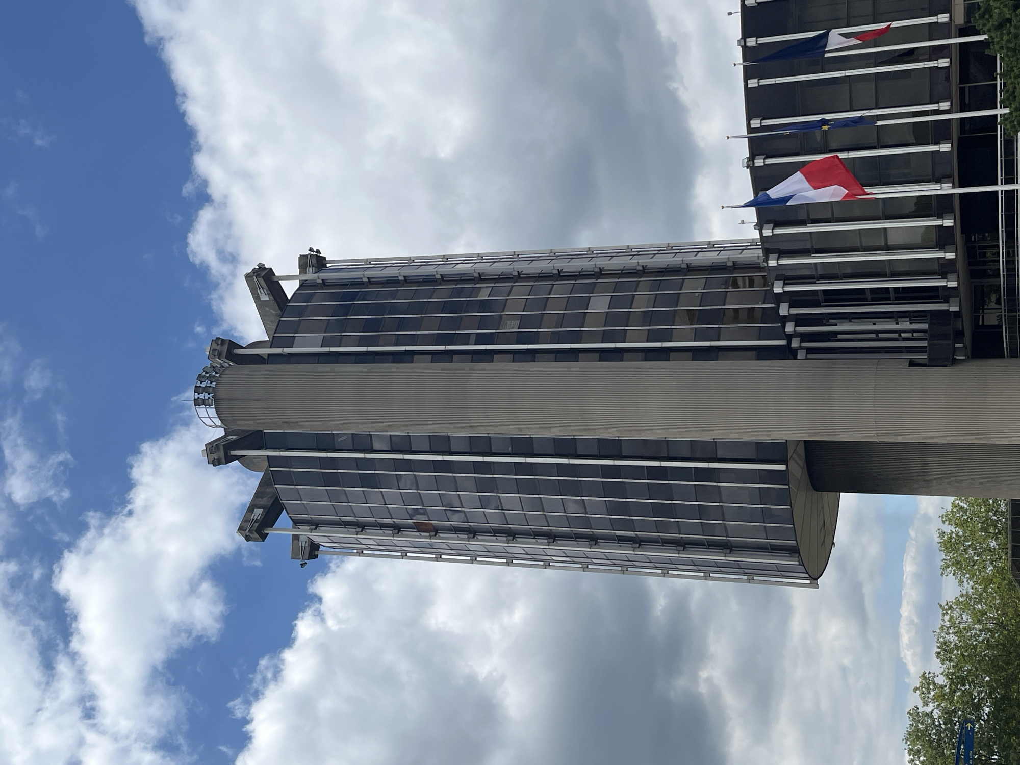 A modern, cylindrical building with a concrete core and a glass facade is visible against a cloudy blue sky. The building has a distinctive flared base and a crown-like structure at its apex. Several French flags and what appears to be an EU flag are flying from flagpoles in front of a lower section of the building, which also features a dark glass facade supported by vertical silver columns. Green trees are visible at the bottom left of the frame. This is likely the Hôtel de Ville (City Hall) of Créteil, France, given its architectural style and the presence of French flags.