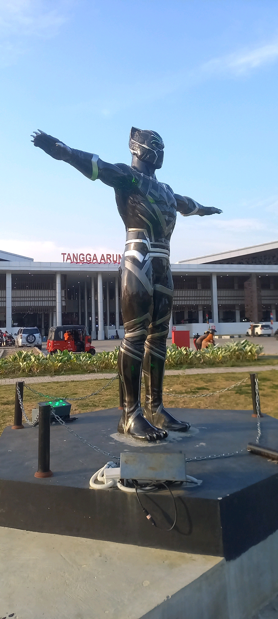 A full-body statue of Black Panther stands in a triumphant pose with arms outstretched in front of a building with "TANGGA ARUN" written in red on its facade. The statue is dark, metallic, and appears to be made of a material with a glossy finish, reflecting the sky and surrounding environment. It is positioned on a dark, rectangular platform. To the left of the statue, a chain link fence separates it from a grassy area where several vehicles are parked, including a white SUV and a red auto-rickshaw. Behind the vehicles and fence, lush green bushes and trees form a border. The building in the background has a modern architectural design with a glass facade and white pillars. The sky above is clear and blue, suggesting a sunny day.