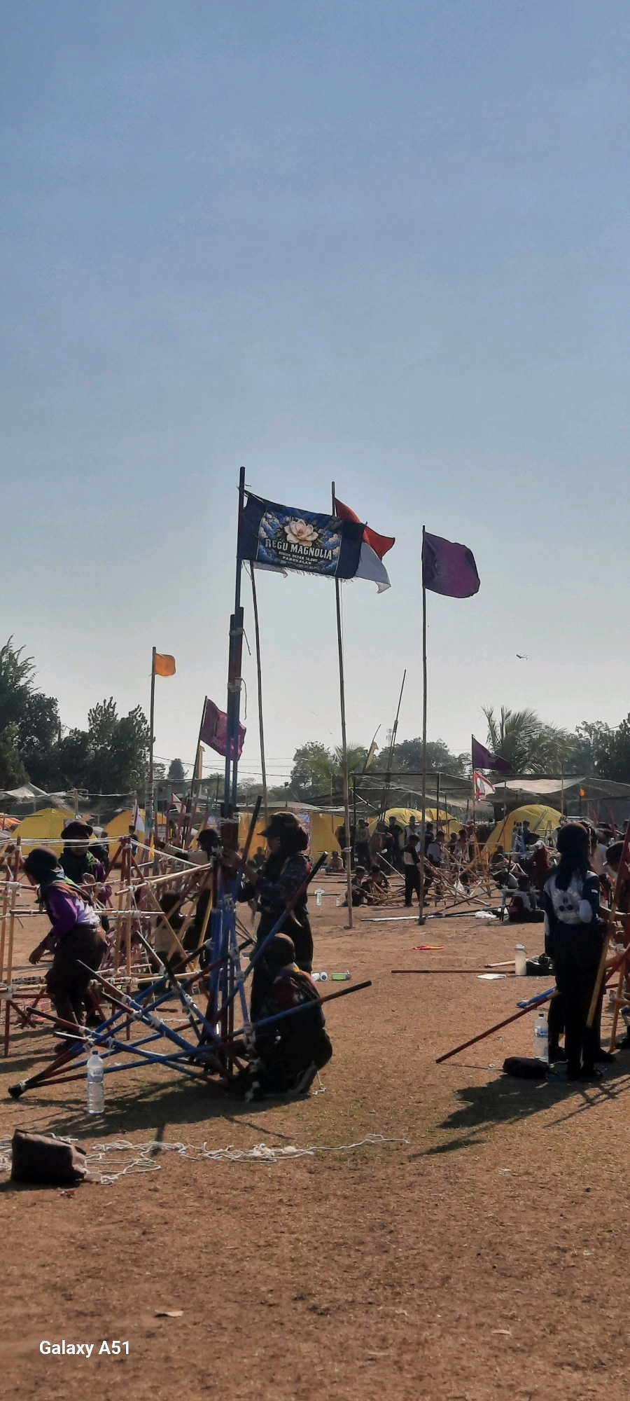In Selopuro, Indonesia, on a clear day with a bright blue sky, a group of scouts participate in an outdoor activity. The scene is set in a dusty, open field with numerous tents erected in the background, suggesting a camp or event. In the foreground, a structure made of blue and red poles, possibly a training apparatus, is prominent. Several scouts, identifiable by their uniforms and hats, are engaged in what appears to be a team activity, some holding poles and seemingly in formation or practicing a skill.
Three flags are visible flying from poles. The most prominent is a dark flag with a lotus flower design and text that reads "REGU MAGNOLIA". Beside it, the Indonesian flag, red and white, is also flying. To the right, a plain purple flag flutters in the breeze. Various scattered objects, including water bottles and bags, are present on the ground. In the distance, more tents and people are visible, indicating a larger gathering. The overall atmosphere suggests a structured outdoor event for young participants.